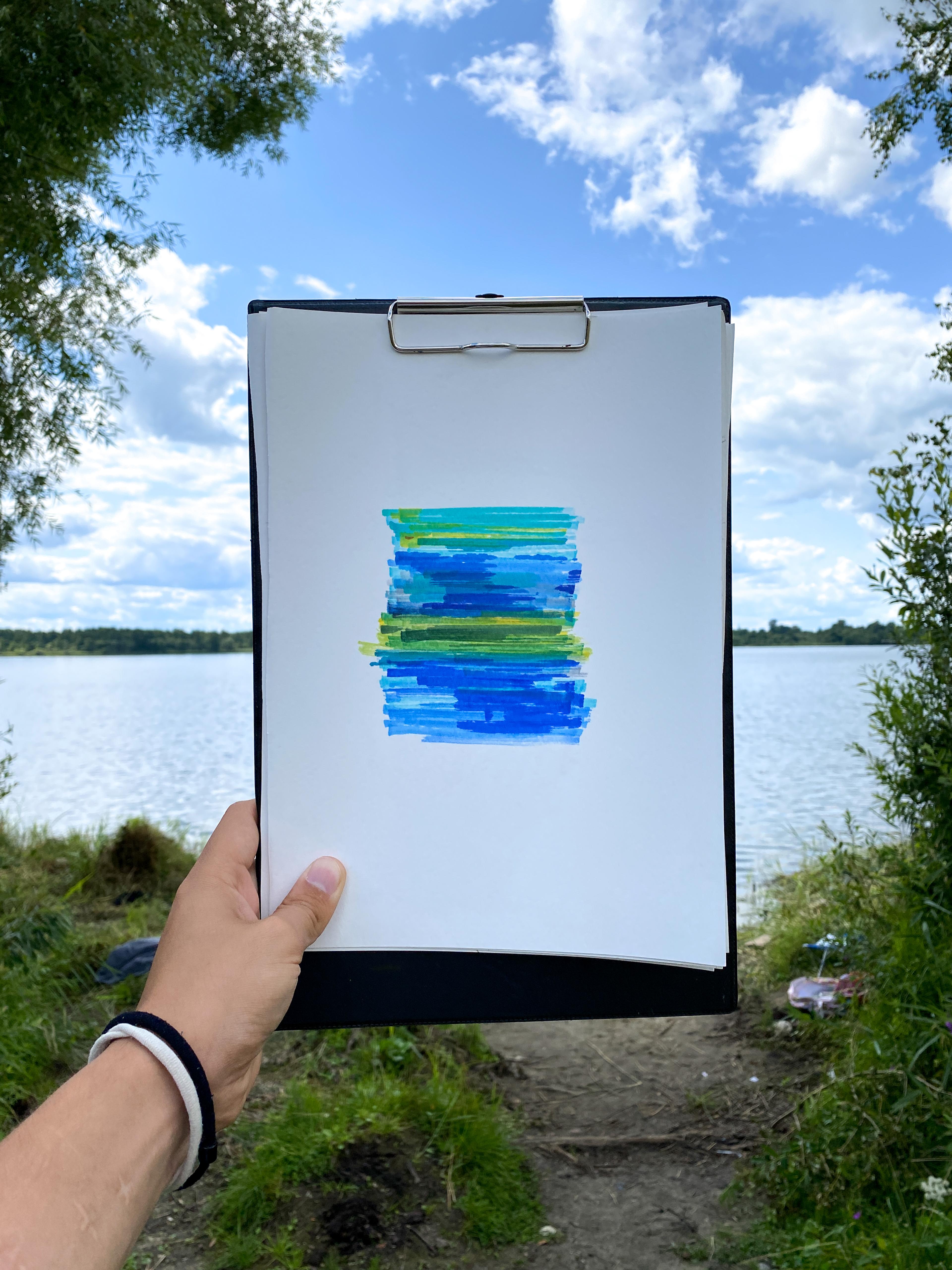 Someone is holding up a small painting of a lake, you can see the lake in the background.