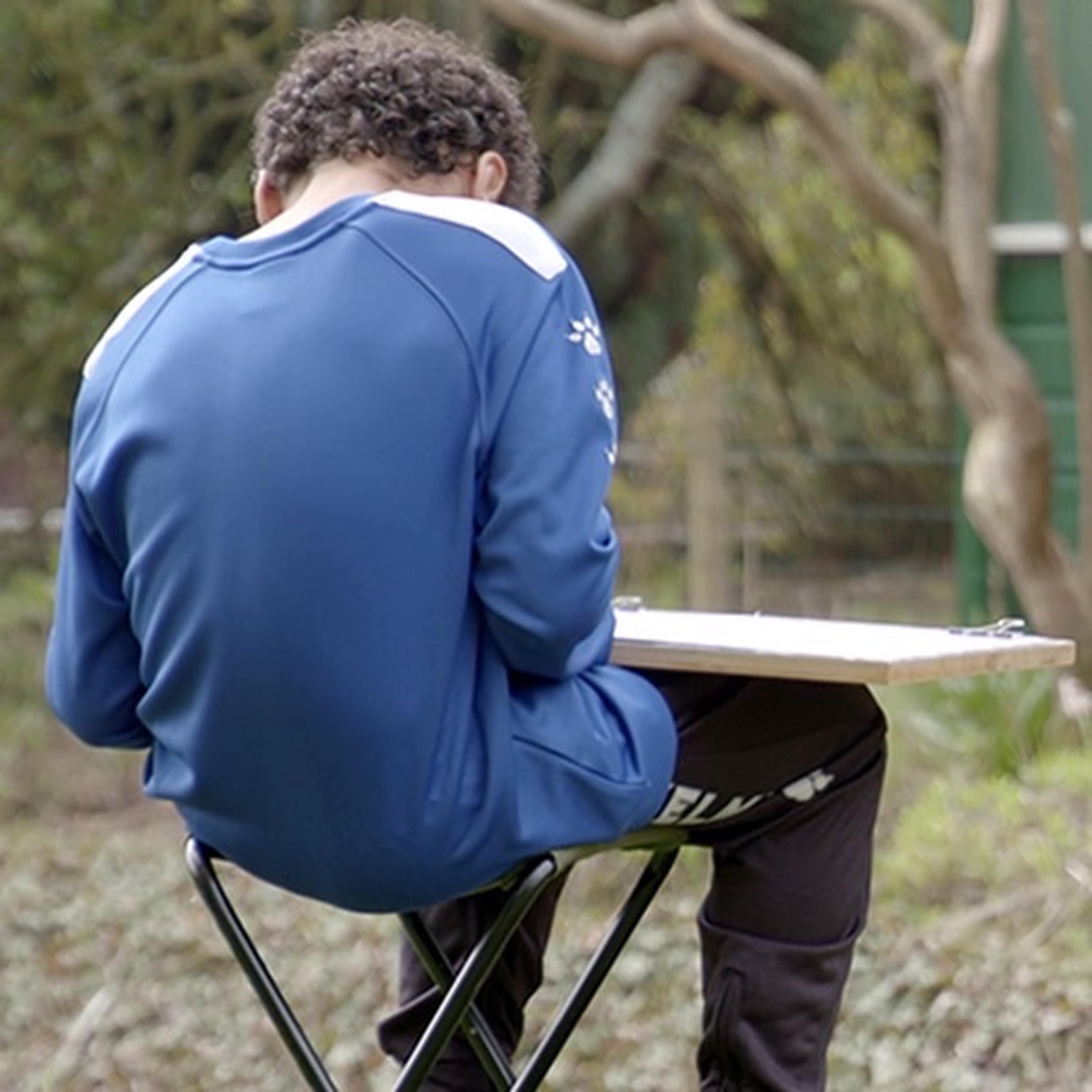 A child with their back to the camera, sat on a folding stool drawing on a piece of paper on a board, in a green, leafy setting.
