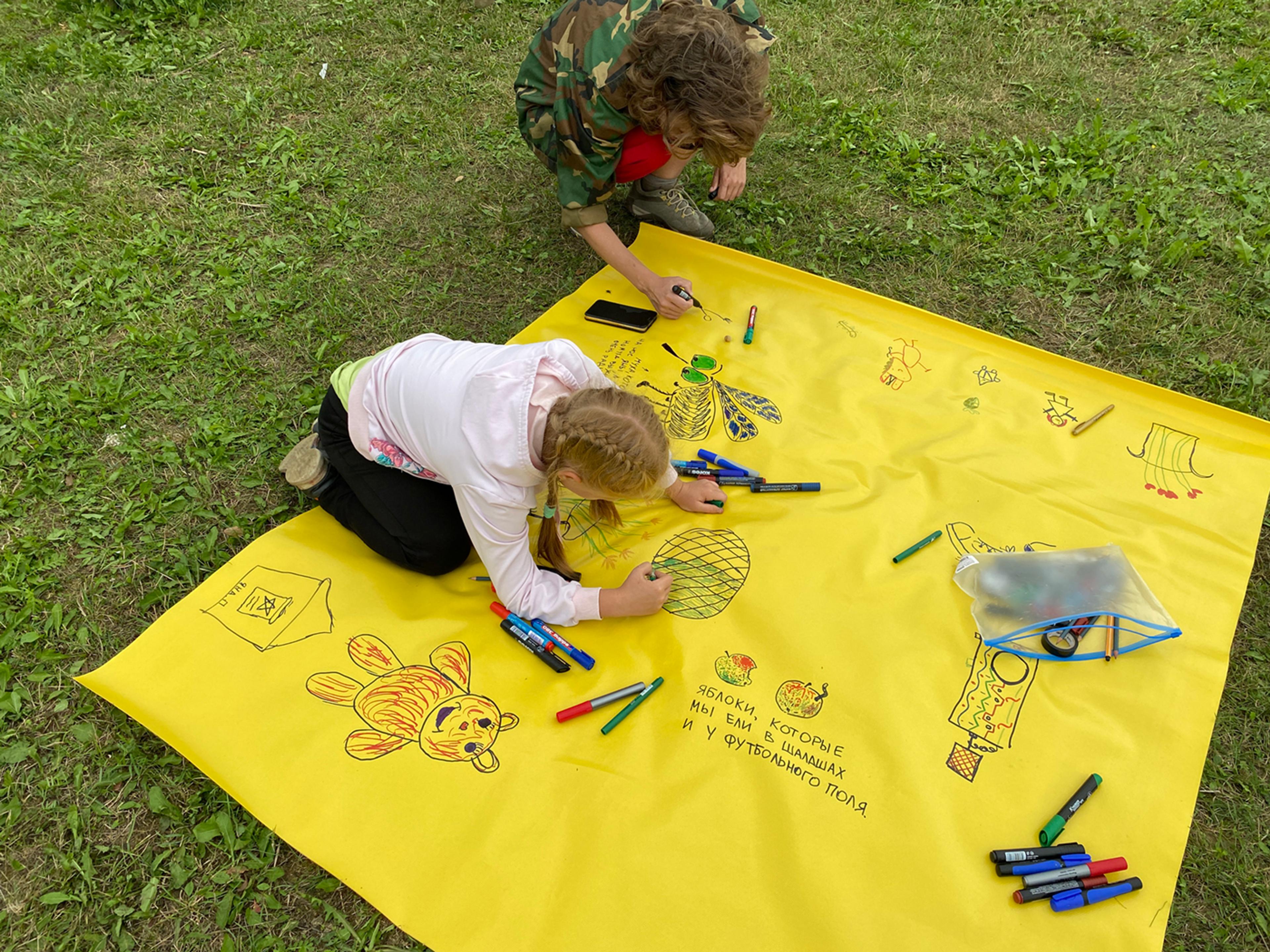 a kid and and an adult drawing with a marker on a big sheet material, that is on the ground