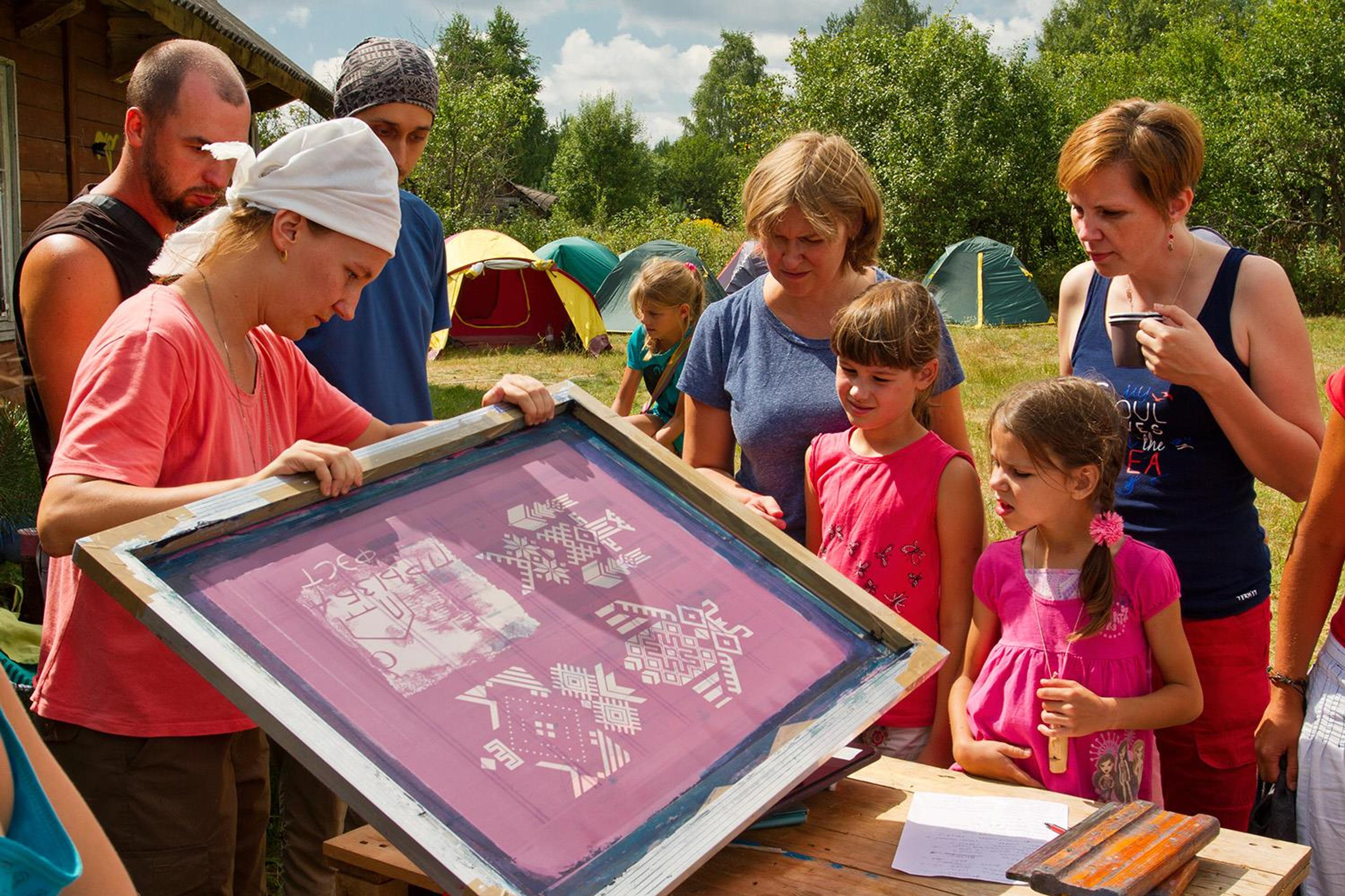 A woman wearing a white headscarf is lifting a screen-printing screen, with a group of people standing around her workshop table.