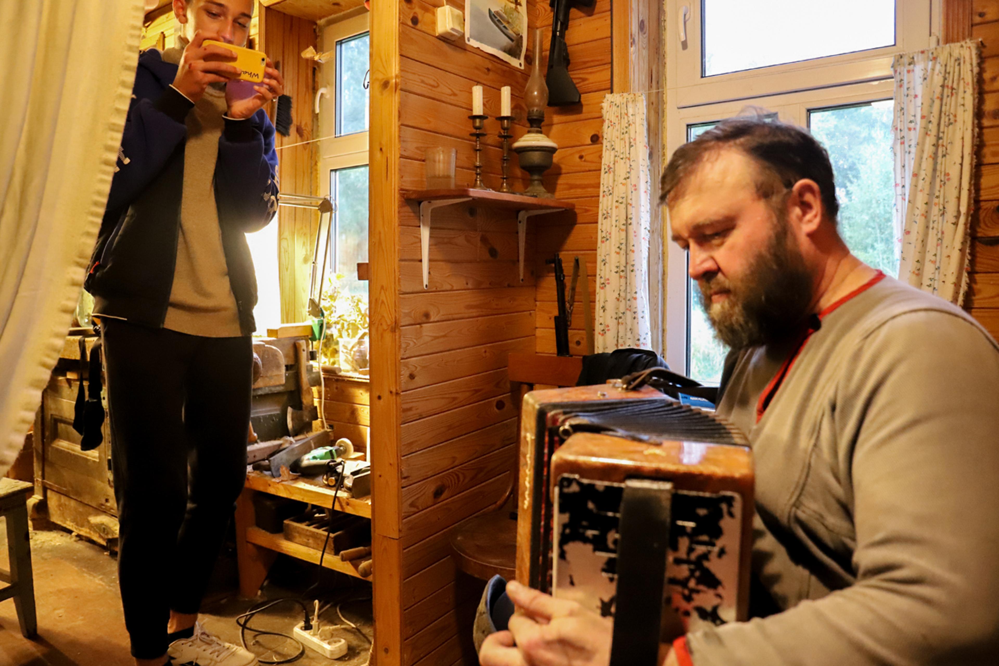 A young men with a camera is filing a bearded man, who is sitting down with a harmonica, playing music in a wooden house.