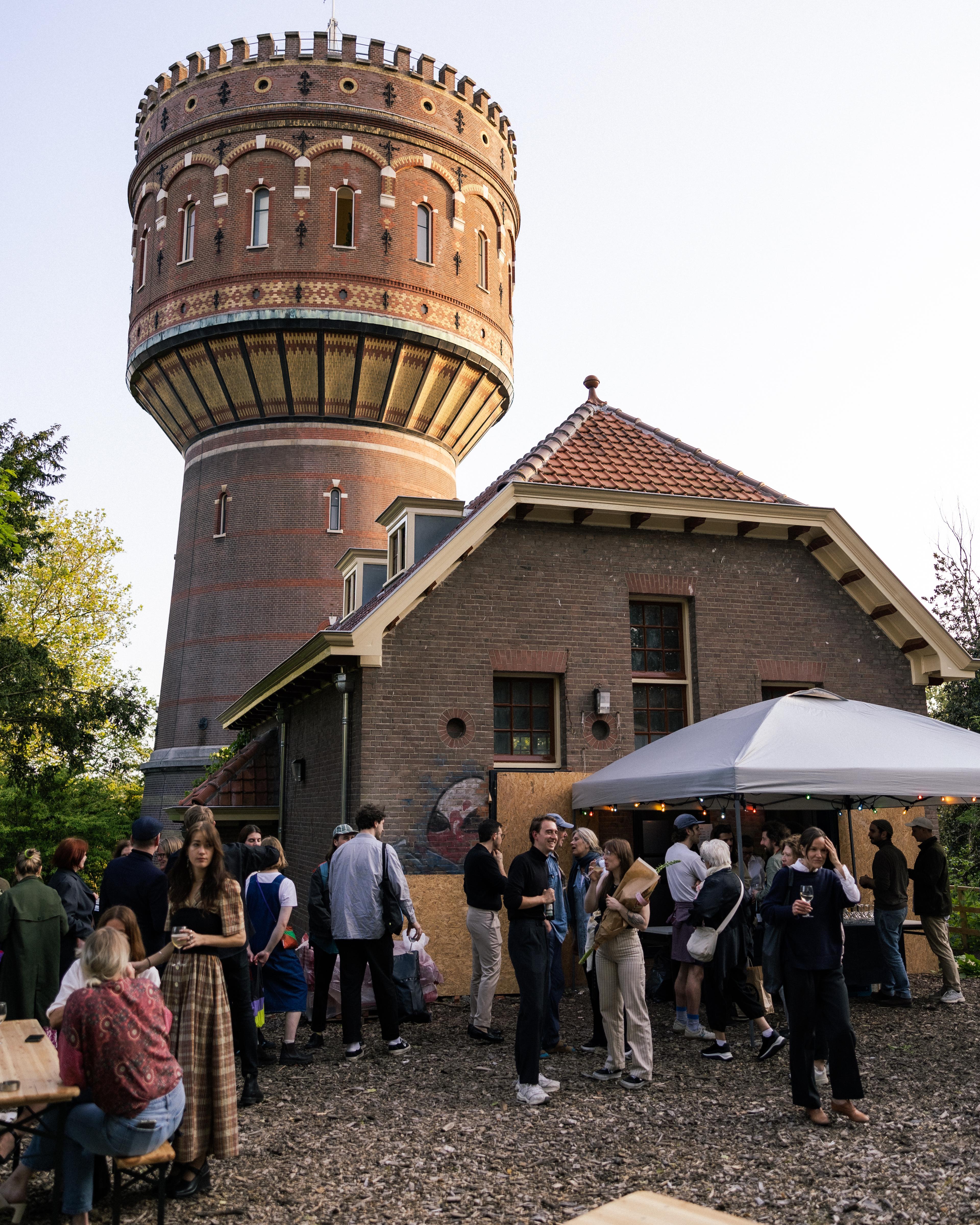 Watertoren in the background, restaurant with outdoor-bar and around 20 poeple standing around the bar with a drink.