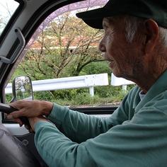 A men in a pale green sweater is driving, and the photo is taken from within the car. You can see green and trees through the car window.