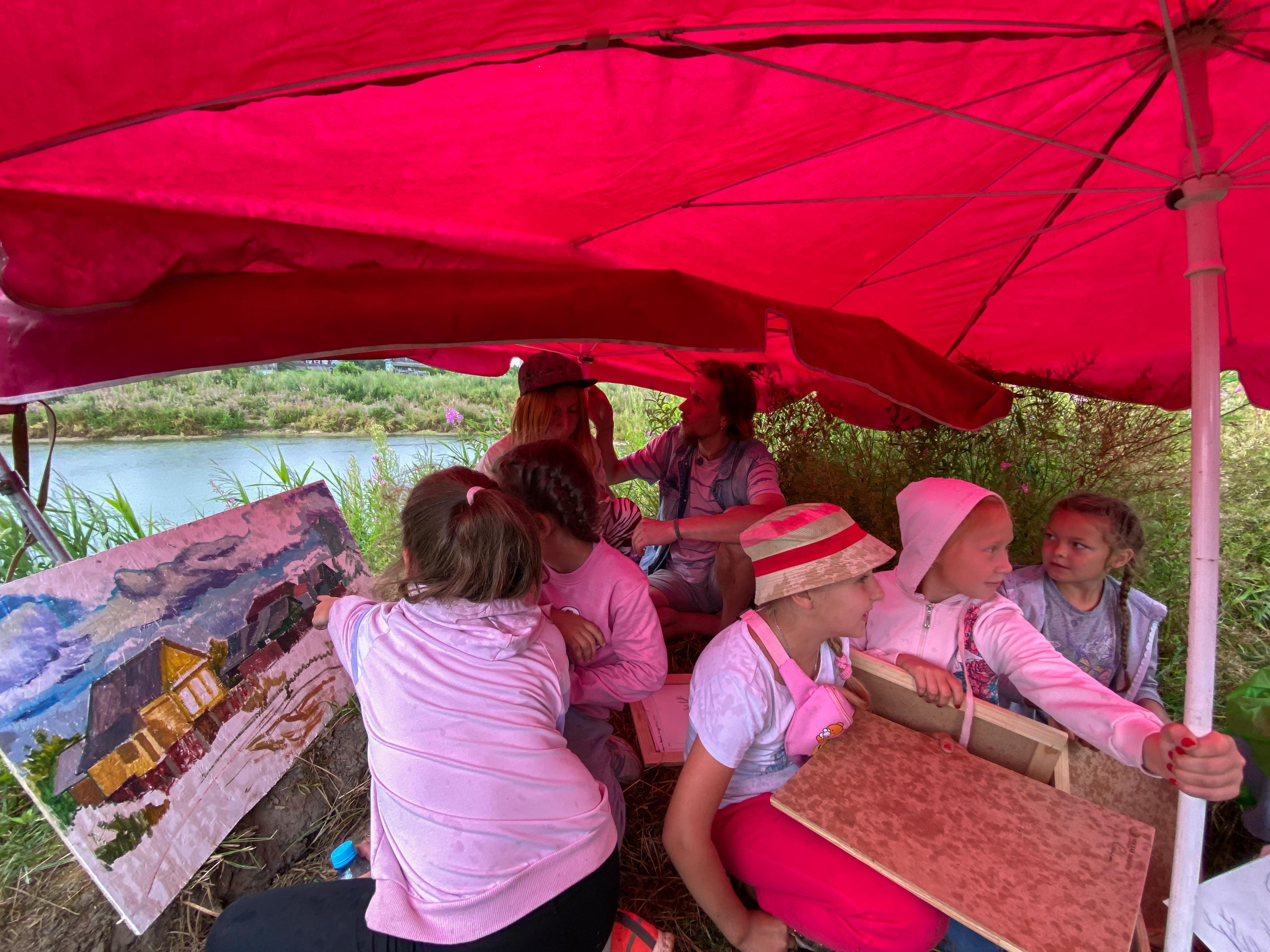 A group of children are sitting outdoors, near a pond and underneath a large red umbrella. They have painting material and on the left is a half finished painting of the landscape. 