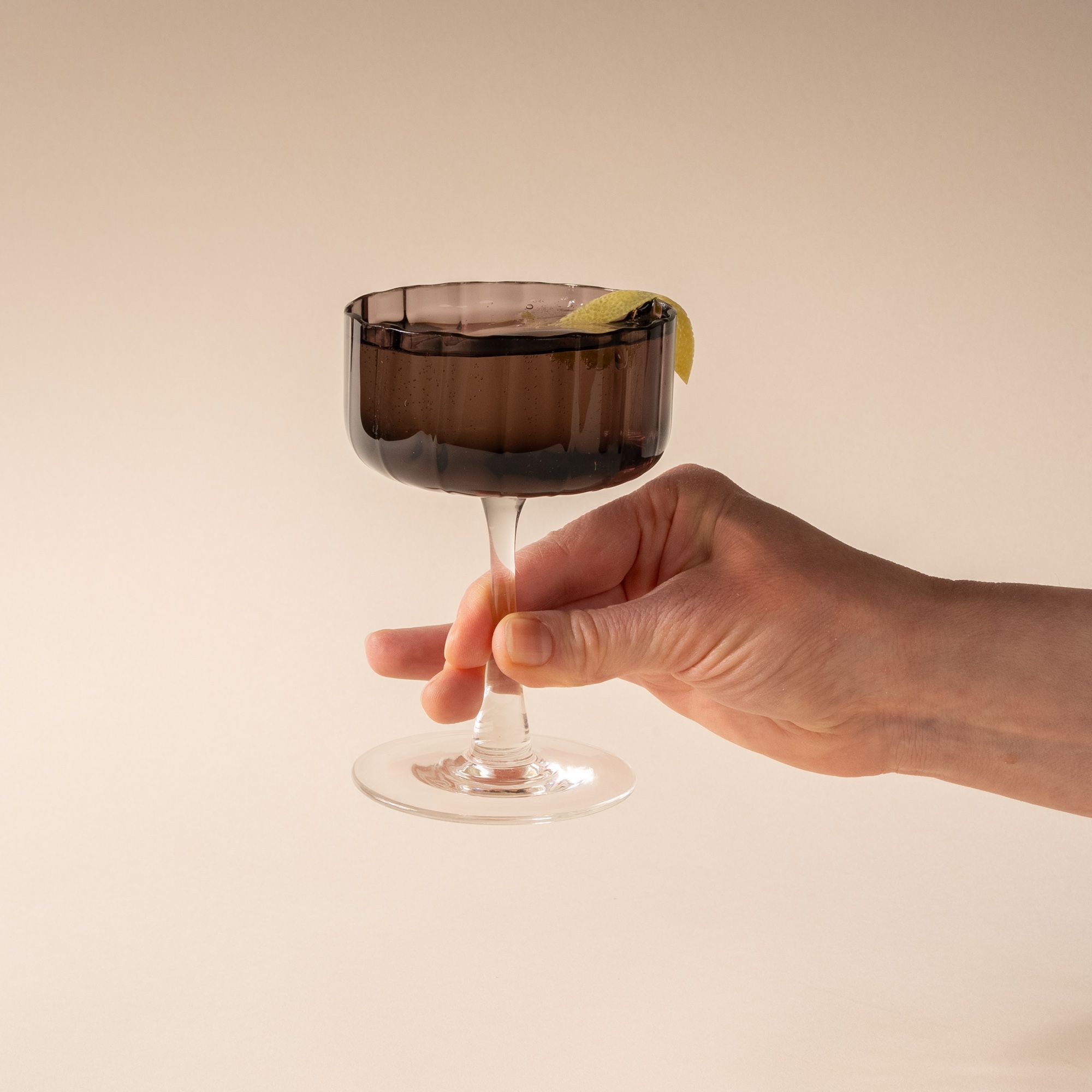 A hand holds a cocktail in a coupe glass with a fluted bowl that is a grey purple color with a clear stem