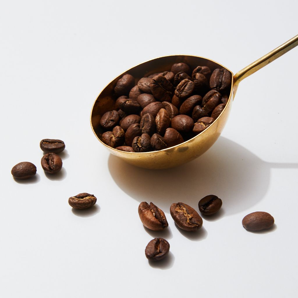 The bowl of a brass coffee scoop filled with coffee beans with more beans in the foreground and part of the scoop's long, thin handle visible