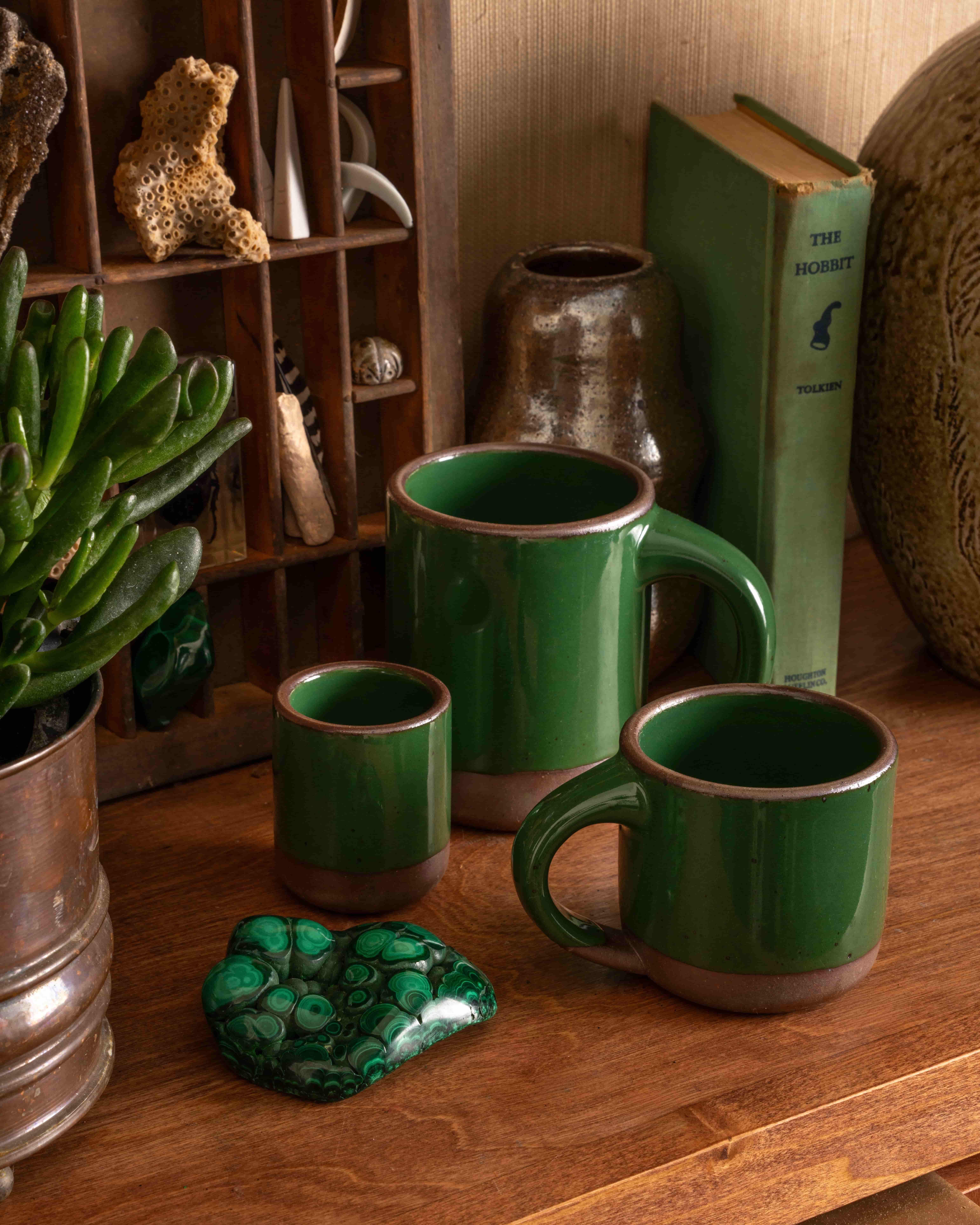 Set of glossy emerald green ceramic mugs on a wooden shelf with malachite stone, plants, pottery, and a copy of The Hobbit.