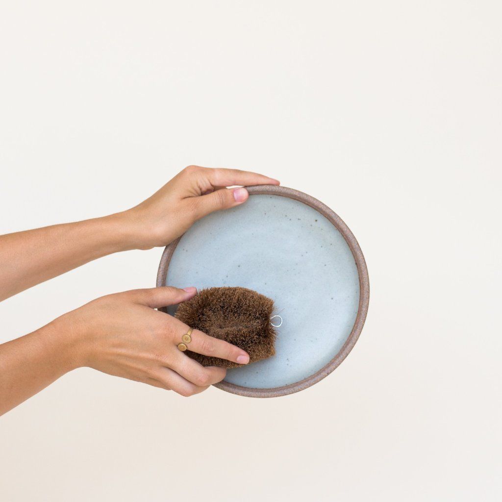 One hand holds the rim of a gray plate while the other hand holds a scrub brush against its surface