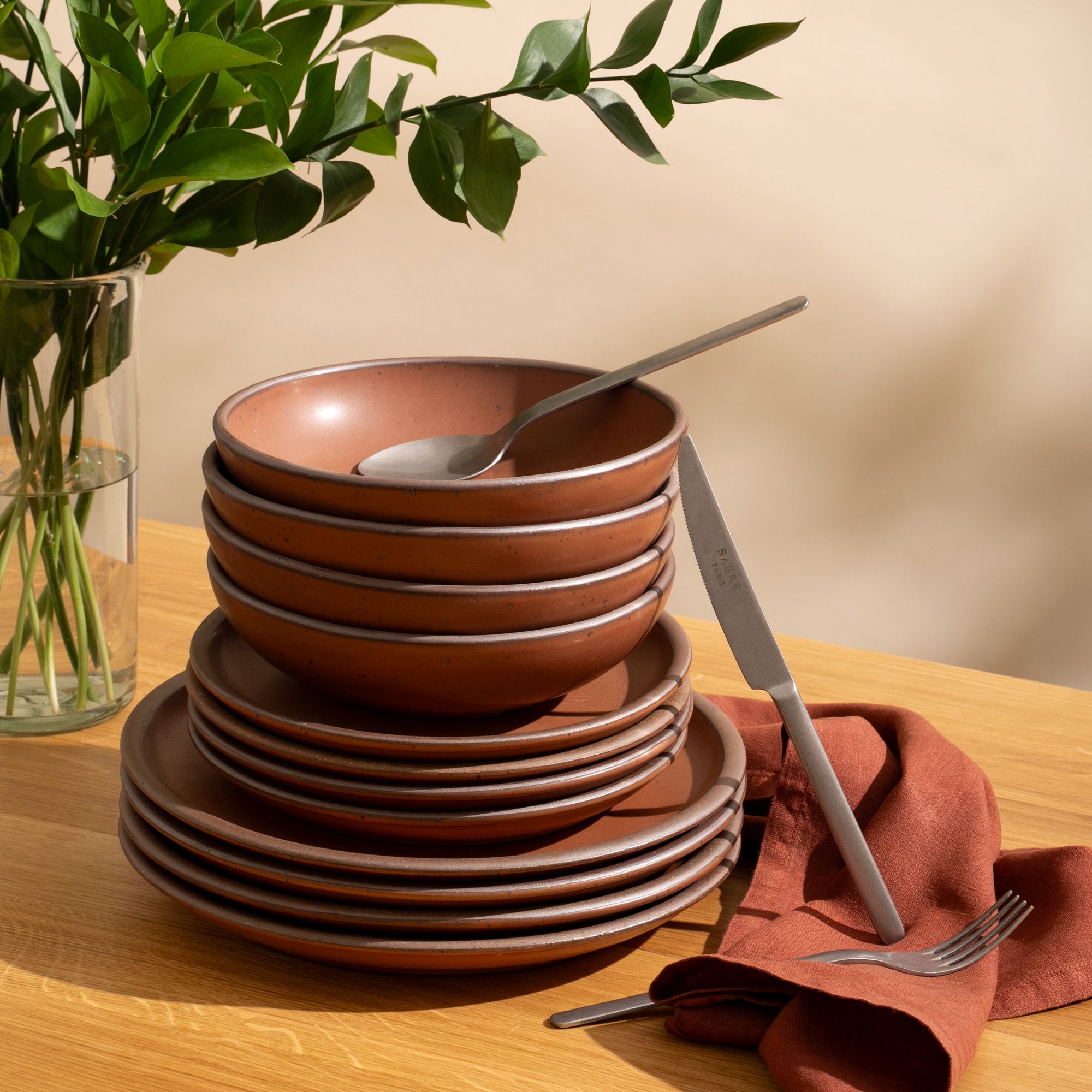 4 ceramic dinner plates, 4 side plates, and 4 shallow dinner bowls in a cool terracotta color with iron speckles and unglazed rims are stacked all together on a table with silverware.