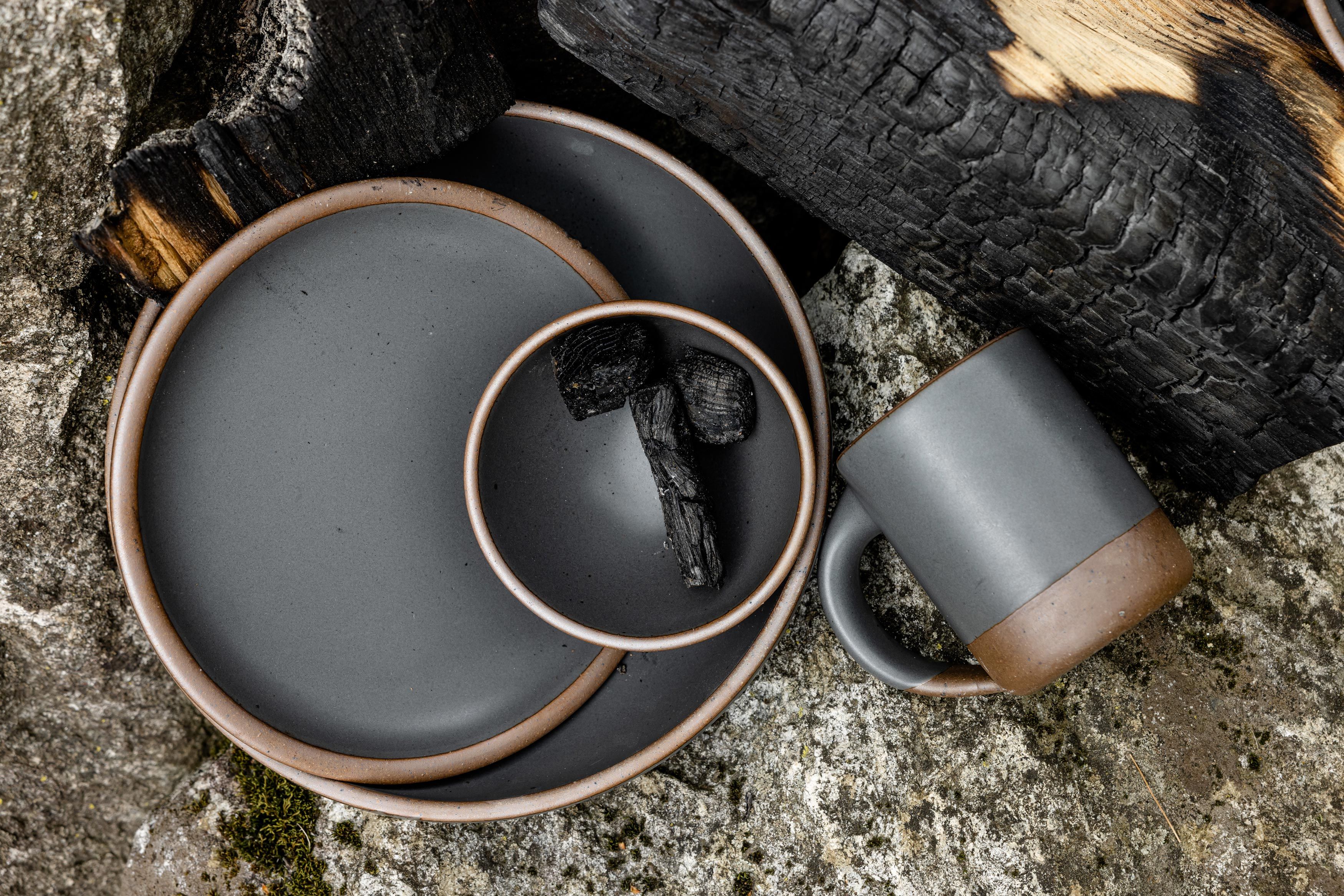 An overhead view of a  stack of grey dinnerware holding charred bits of firewood and a mug on its side lying on a rock amongst large piece of partially charred firewood.