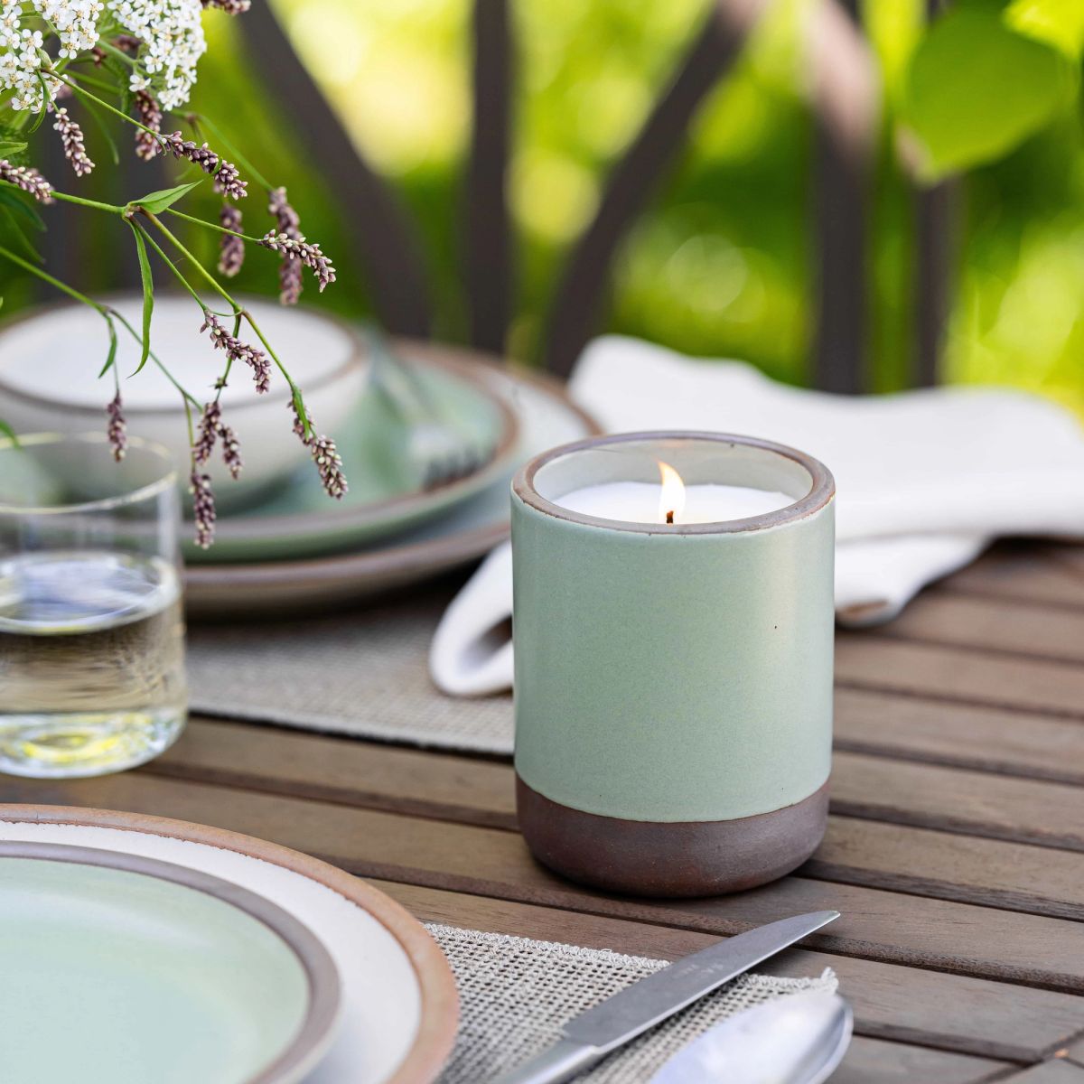 On an outdoor table, there is a few place settings and an outdoor candle in a ceramic cylindrical vessel in a sage green color that is lit.