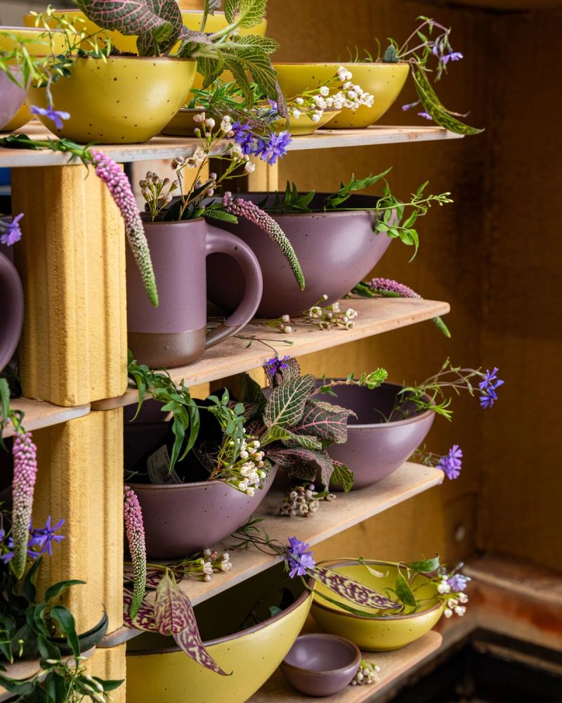 A kiln cart loaded with ceramic mugs and bowls in zesty yellow and soft mauve purple colors, with flowers.