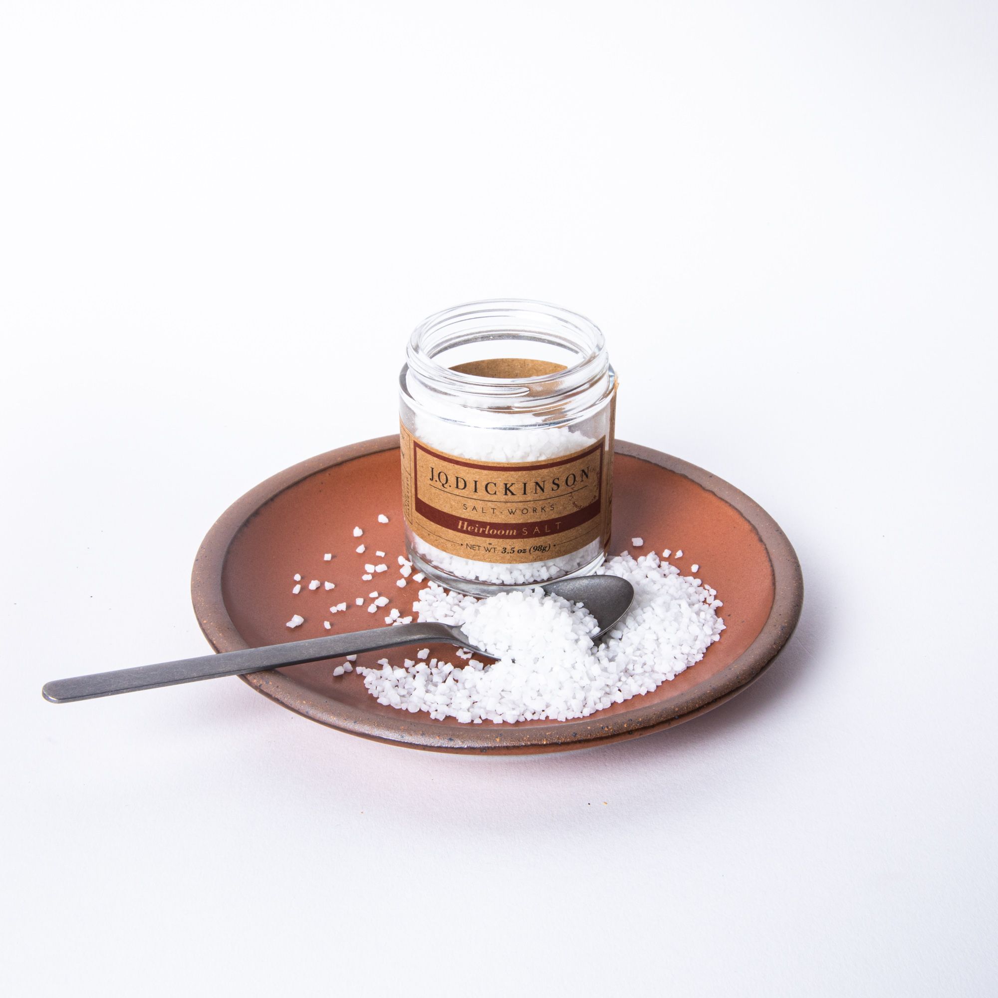 A short open jar filled with salt with a kraft brown label. The jar sits on a plate with salt and a spoon