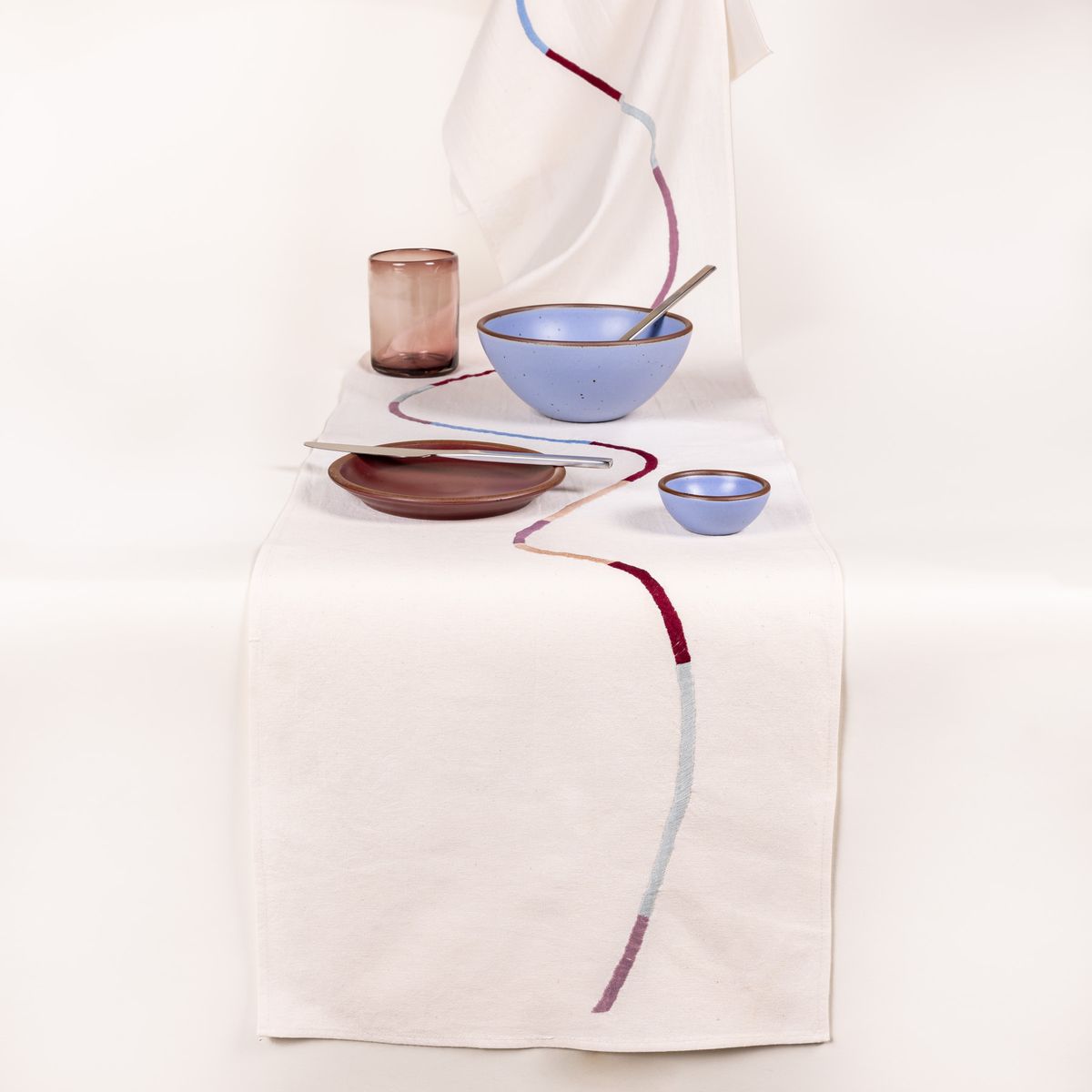 Ceramic dishes and a glass sit on a table runner in a natural cotton with a thin embroidered line curving down the runner in color blocks of purple, peach, and periwinkle colors