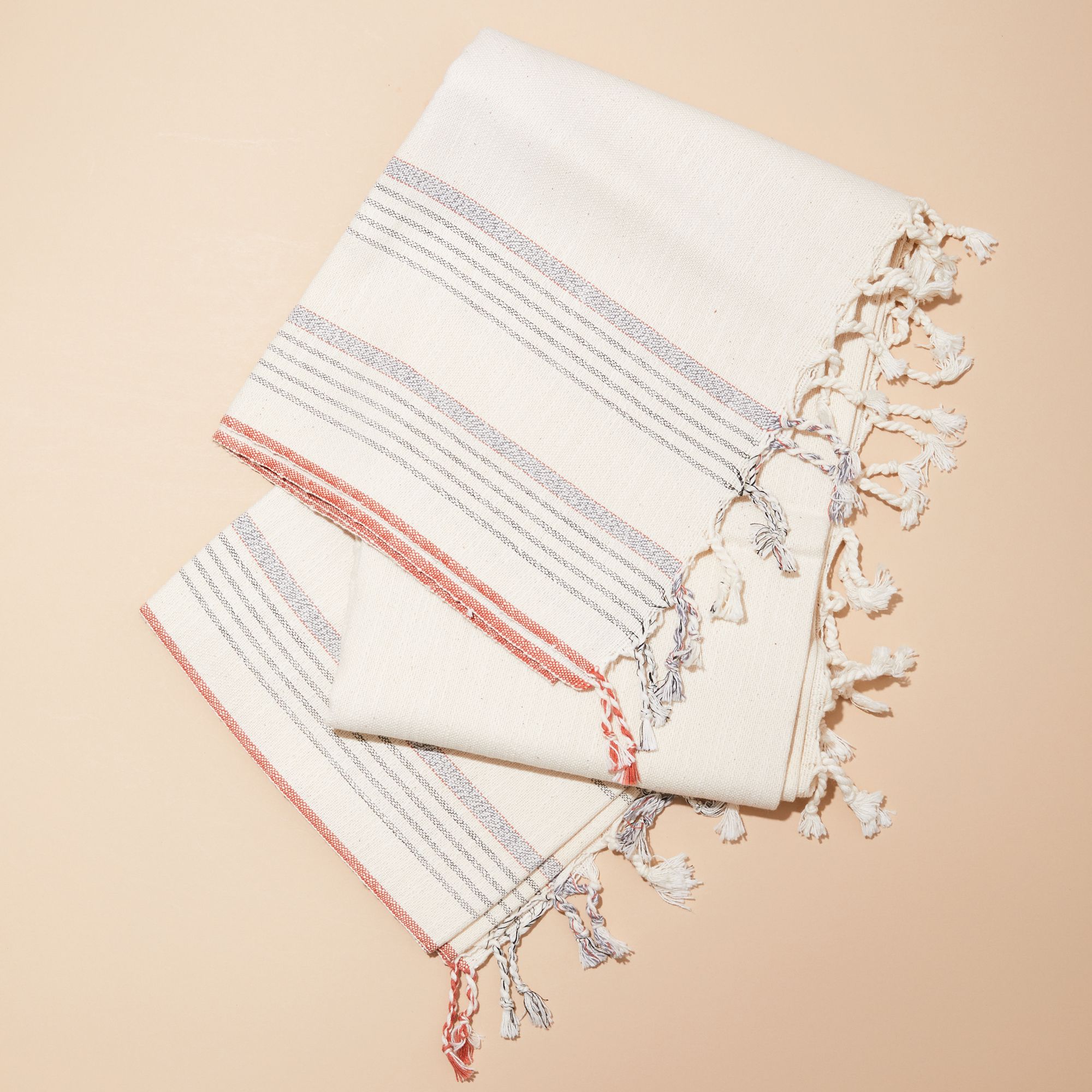 A cream-colored cotton Turkish towel with blue and coral stripes and tassels 