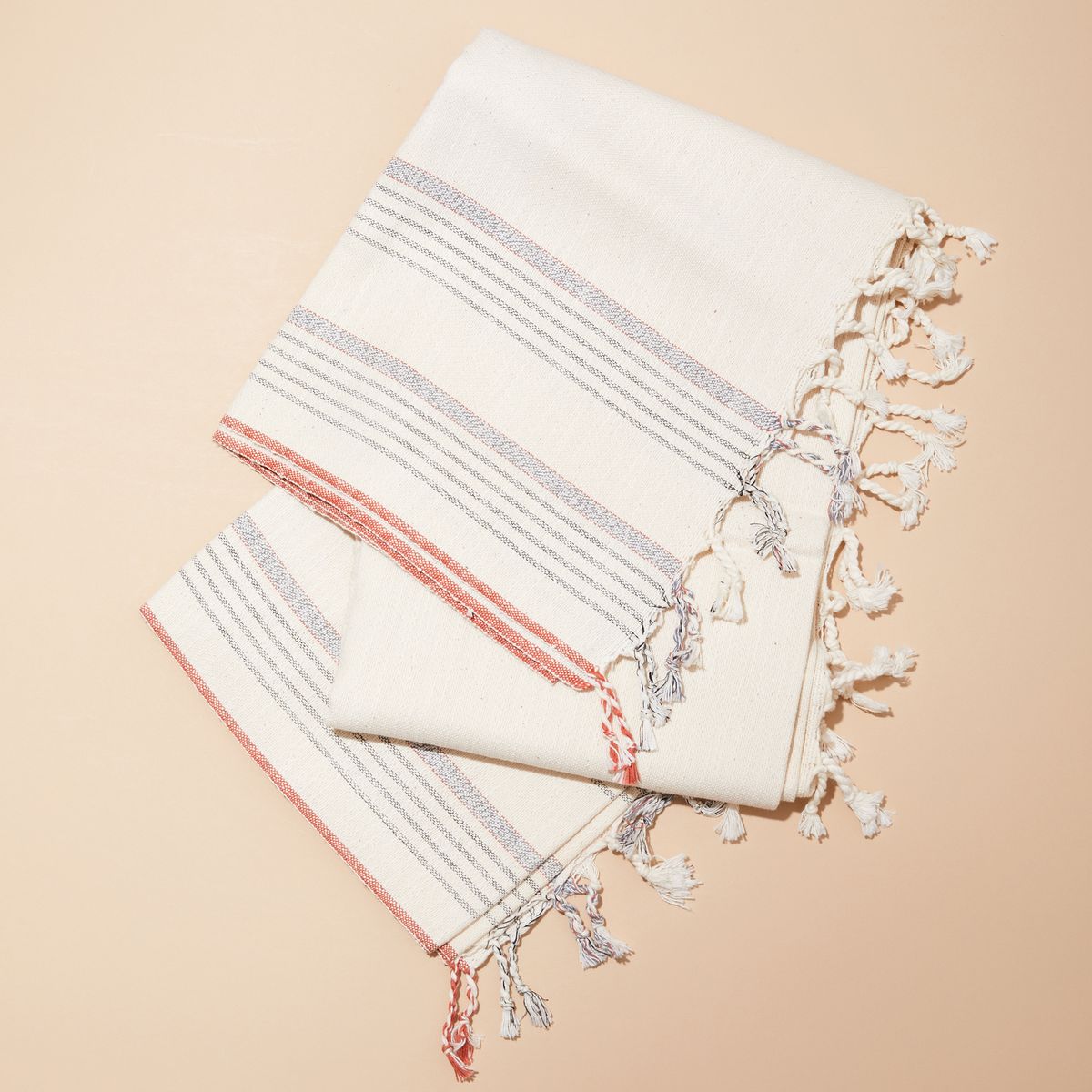 A cream-colored cotton Turkish towel with blue and coral stripes and tassels