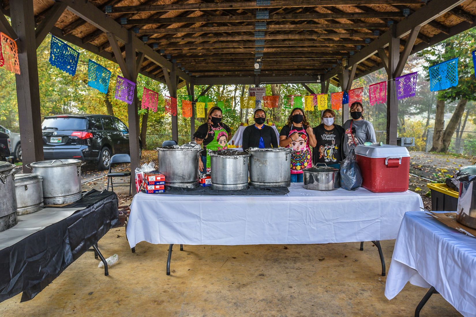 Outside under a pavillion, 5 people are gathered behind a table ready to serve food from large pots and a cooler