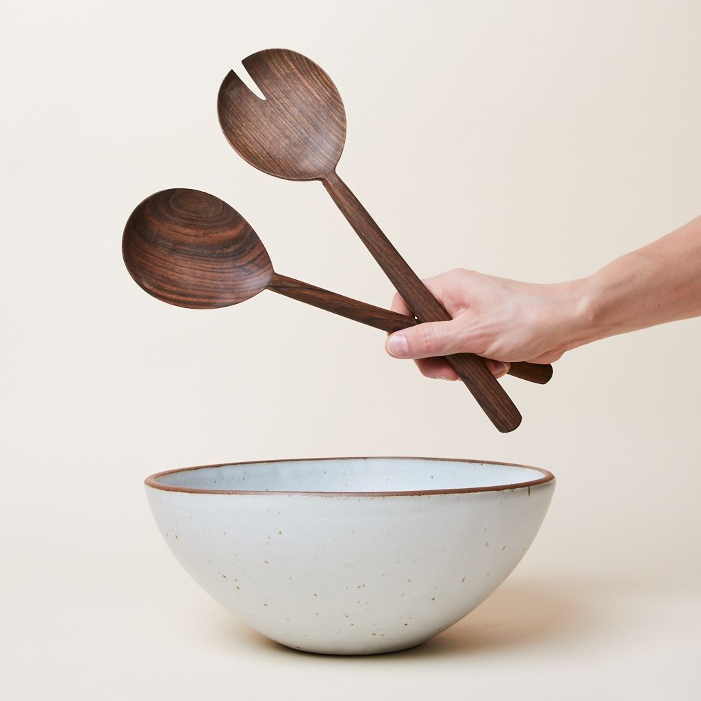 Above an East Fork Soup Bowl in Eggshell, a hand holds a set of two walnut serving spoons, one with a notch cut out