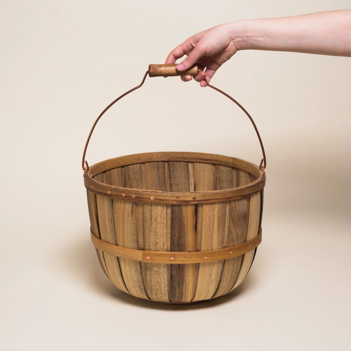 A hand holding the handle of a large tall round rustic wooden basket with a metal and wood handle