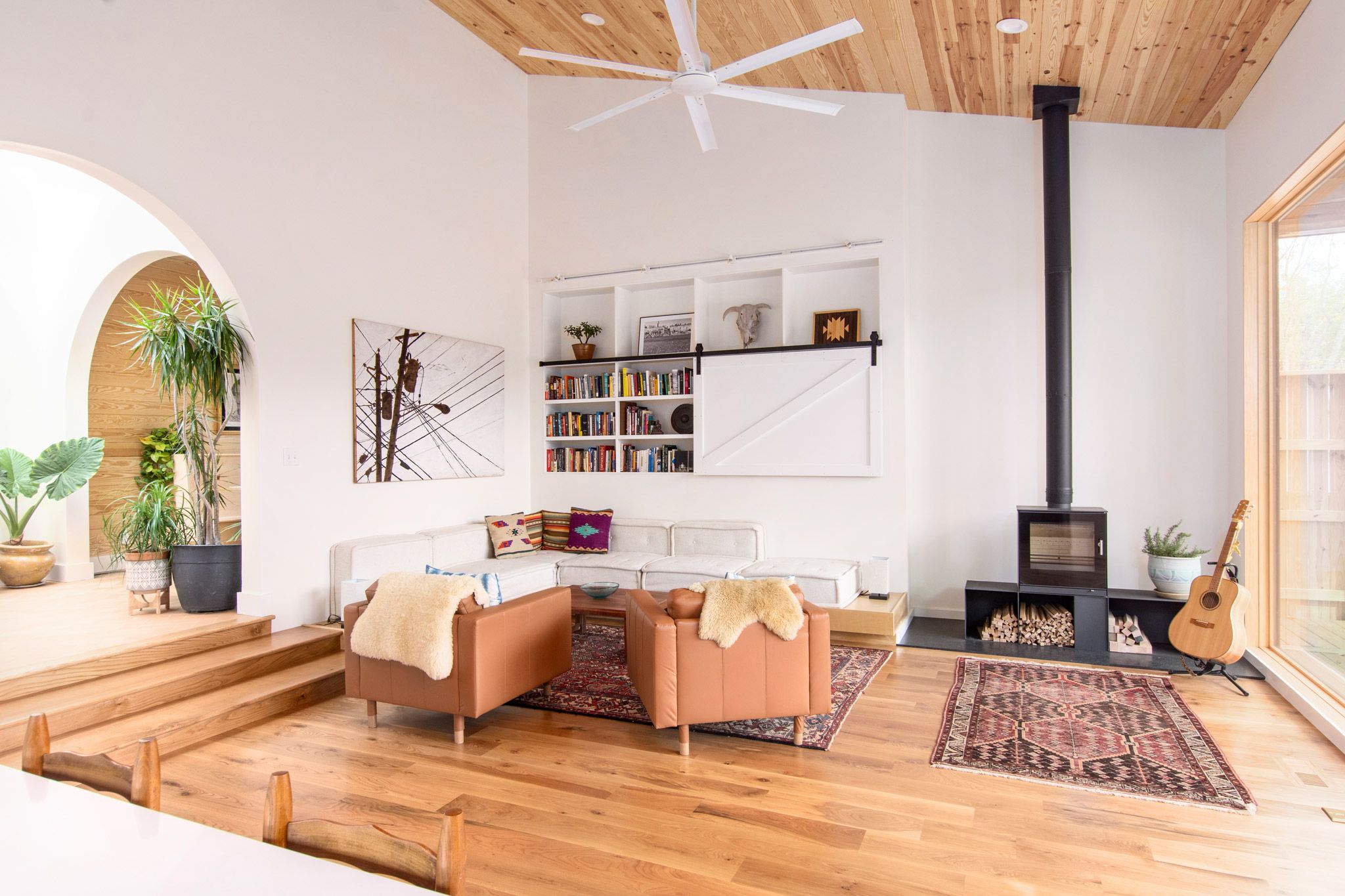 A living room featuring a large white sectional couch, minimal black furnace, brown leather chairs, potted plants, and a large window.