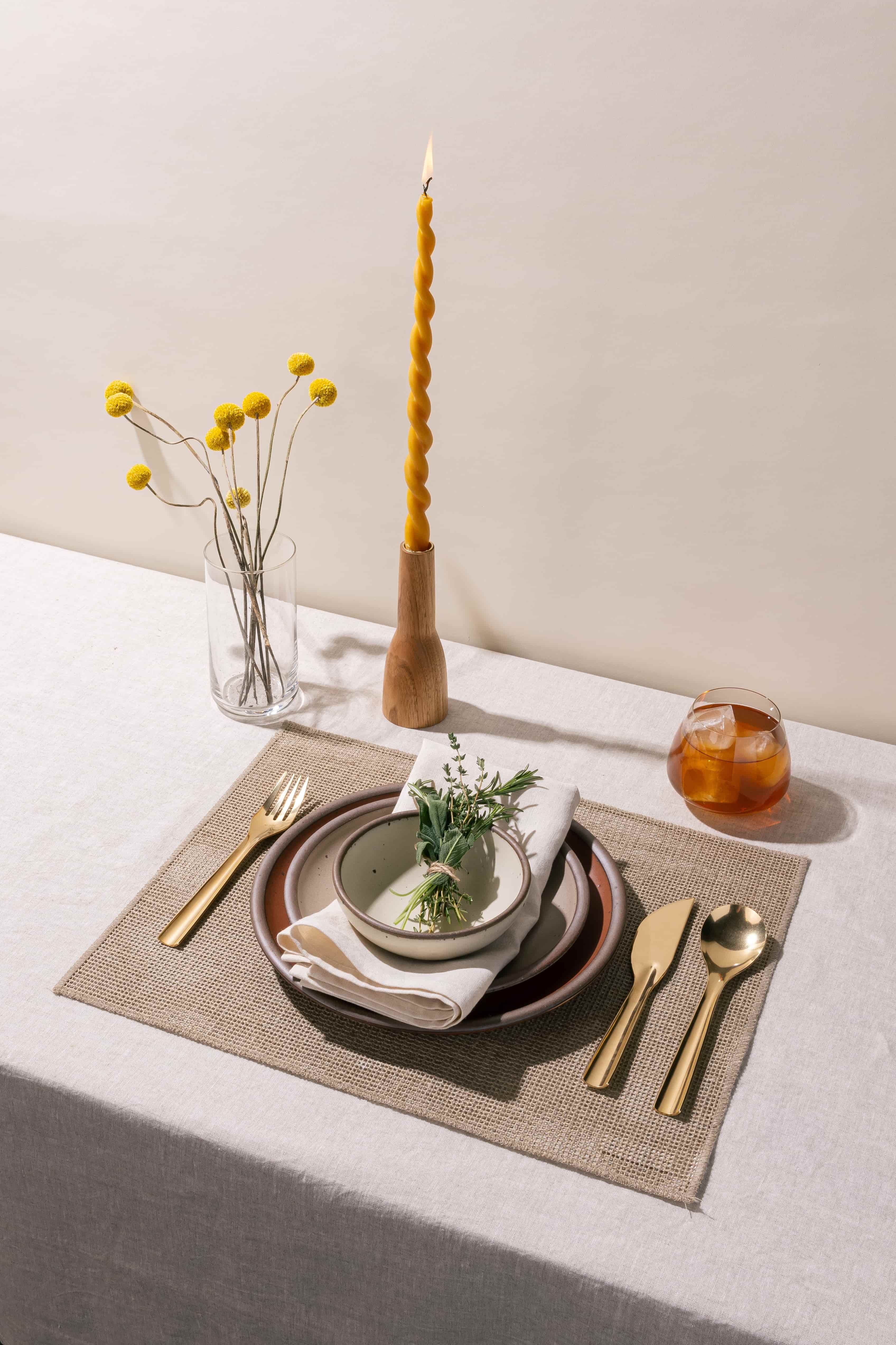 A table setting featuring ceramic plates in terracotta and neutral colors, brass flatware, a whiskey glass, and a twisted taper candle.