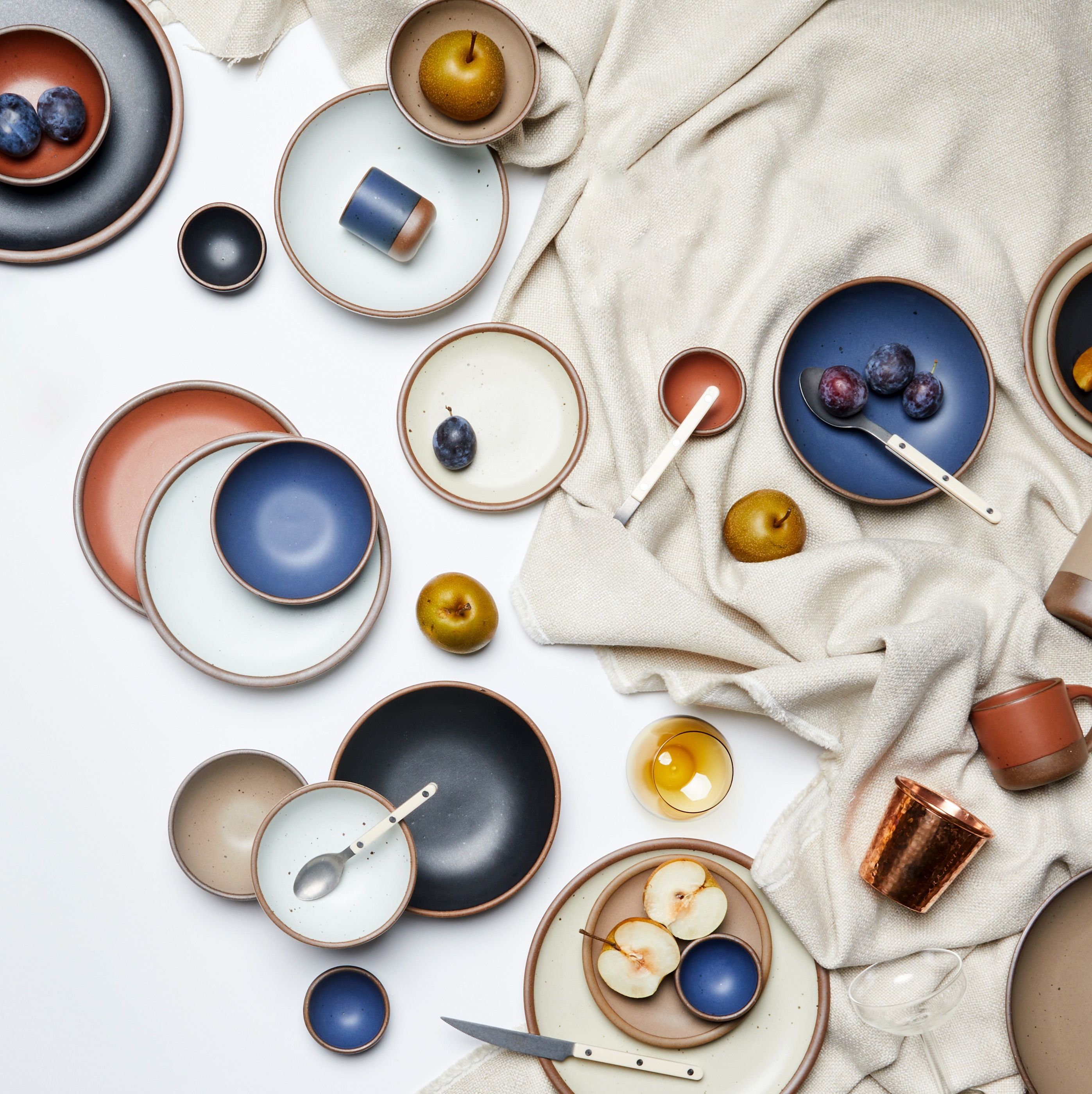 An overhead view of artfully arranged ceramic plates in neutral, blue, and terracotta colors, surrounded by various flatware, pears, glassware and more. All are artfully arranged with a white background and neutral fabric.