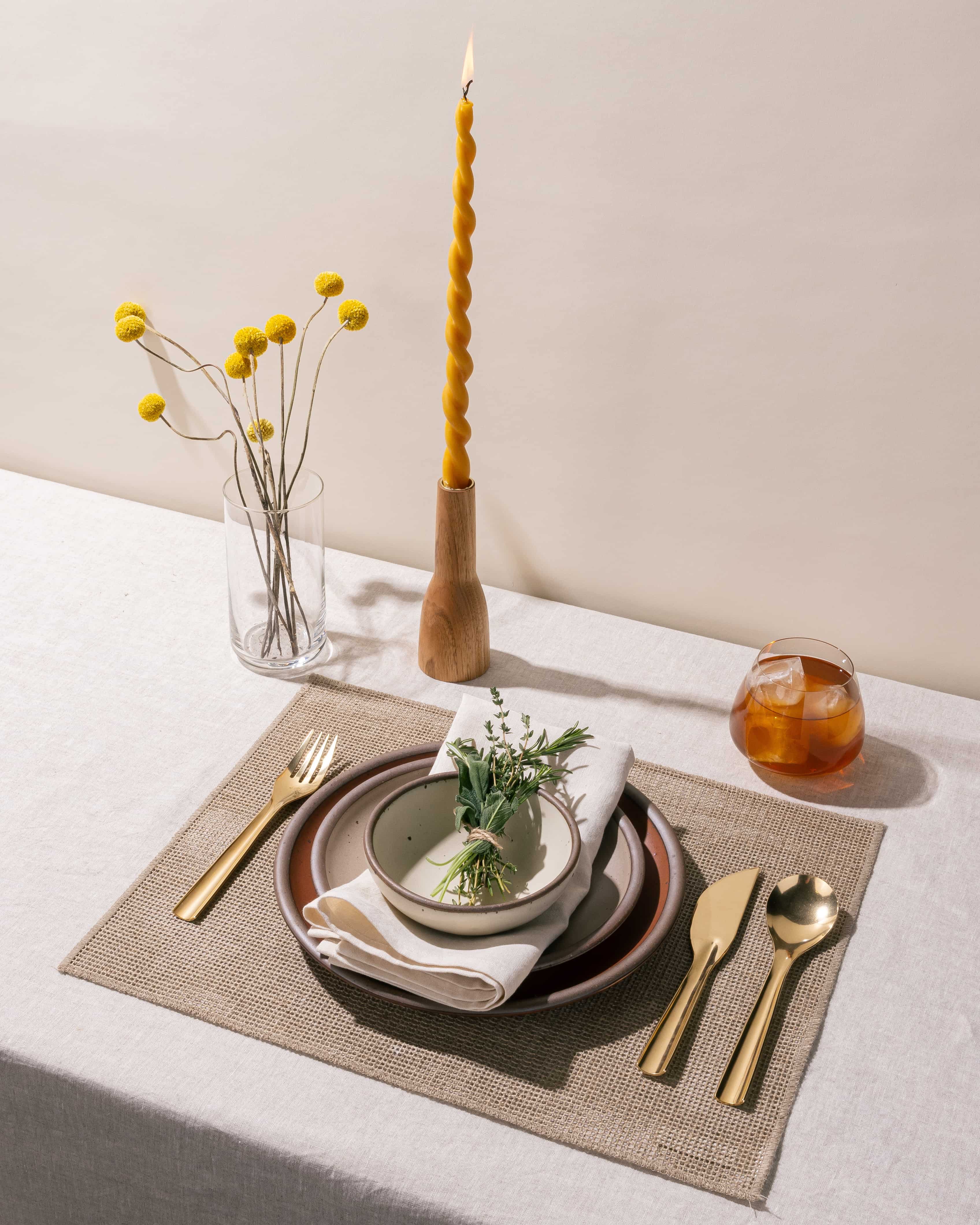 A table setting featuring a stack of ceramic plates, brass flatware, drink, taper candle, and more.