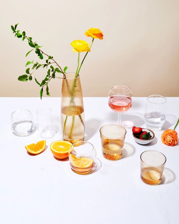 Table with various glasses filled with water in different shapes, forms and sizes. Table is also adorned with fruit, a carafe with flower arrangement, and a small bowl of fruit.