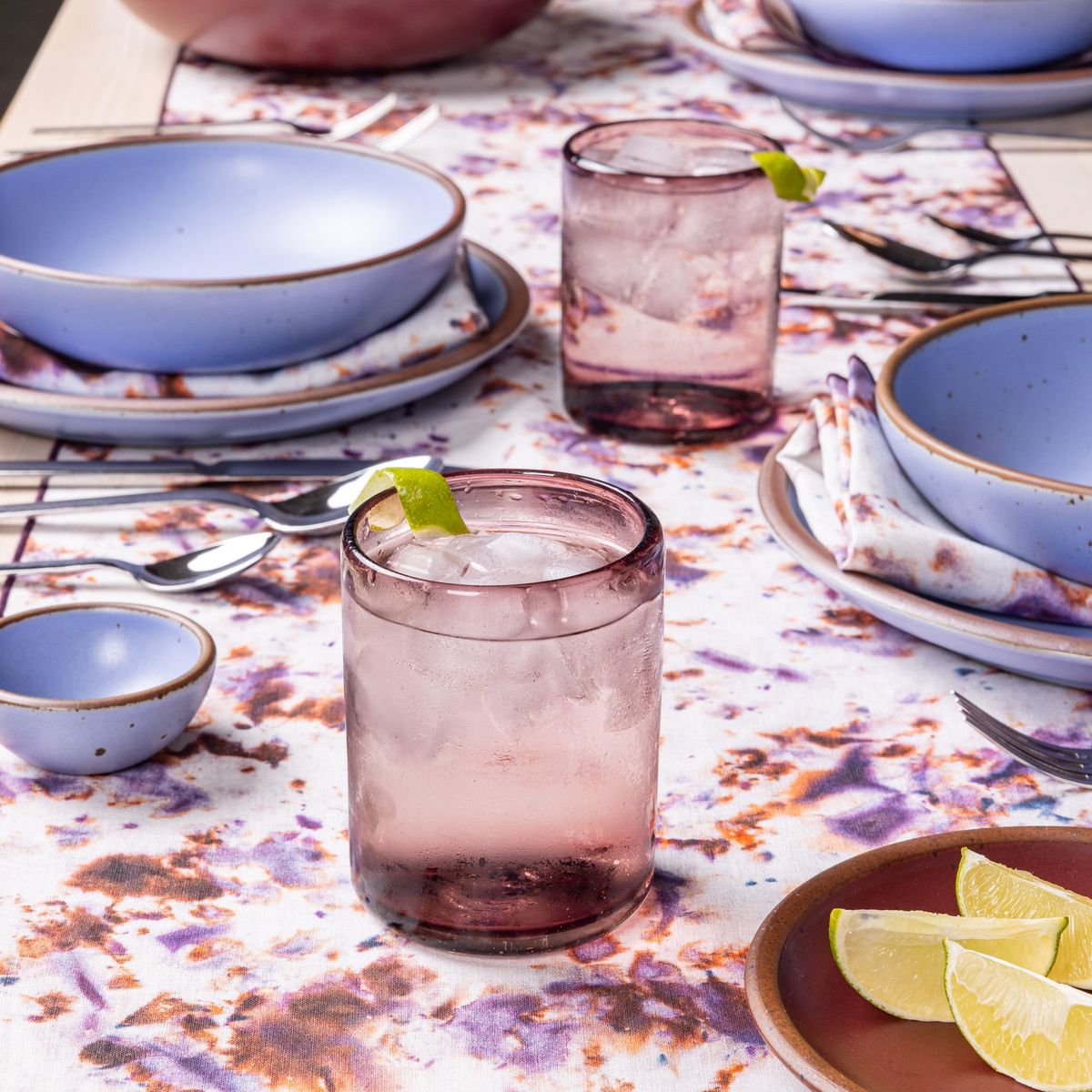 On a table, are 2 light plum short drinking glasses filled with a drink surrounded by periwinkle ceramic plates and bowls