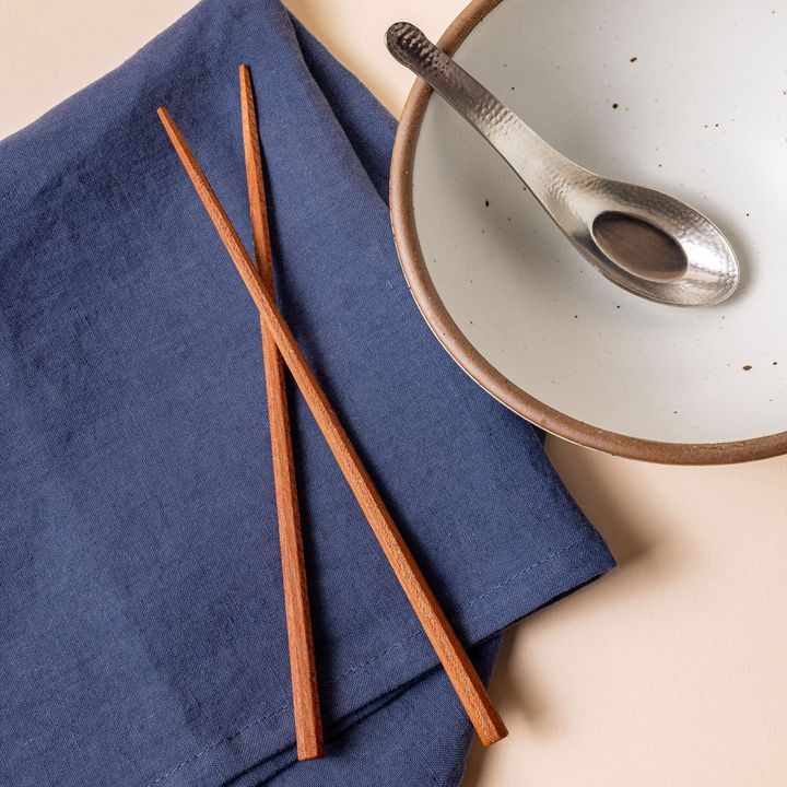 Simple light brown chopsticks laying on a muted blue napkin next to a soup bowl with a steel soup spoon inside.