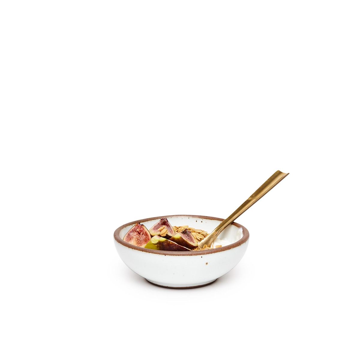 A small shallow ceramic bowl in a cool white color featuring iron speckles and an unglazed rim, filled with granola and a brass spoon