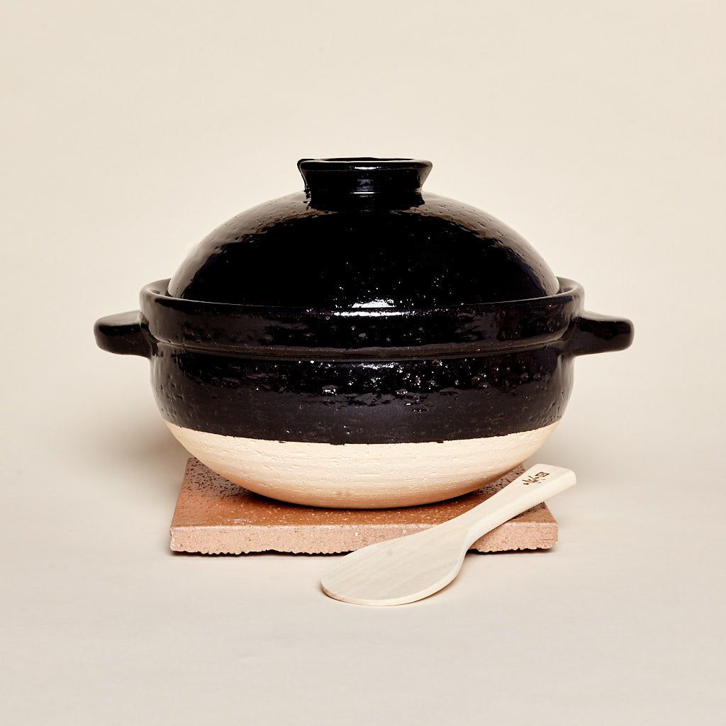 A black-and-neutral color claypot with handles and a shallow-dome shaped lid. The cooking vessel sits atop a wooden board next to a wooden spoon.