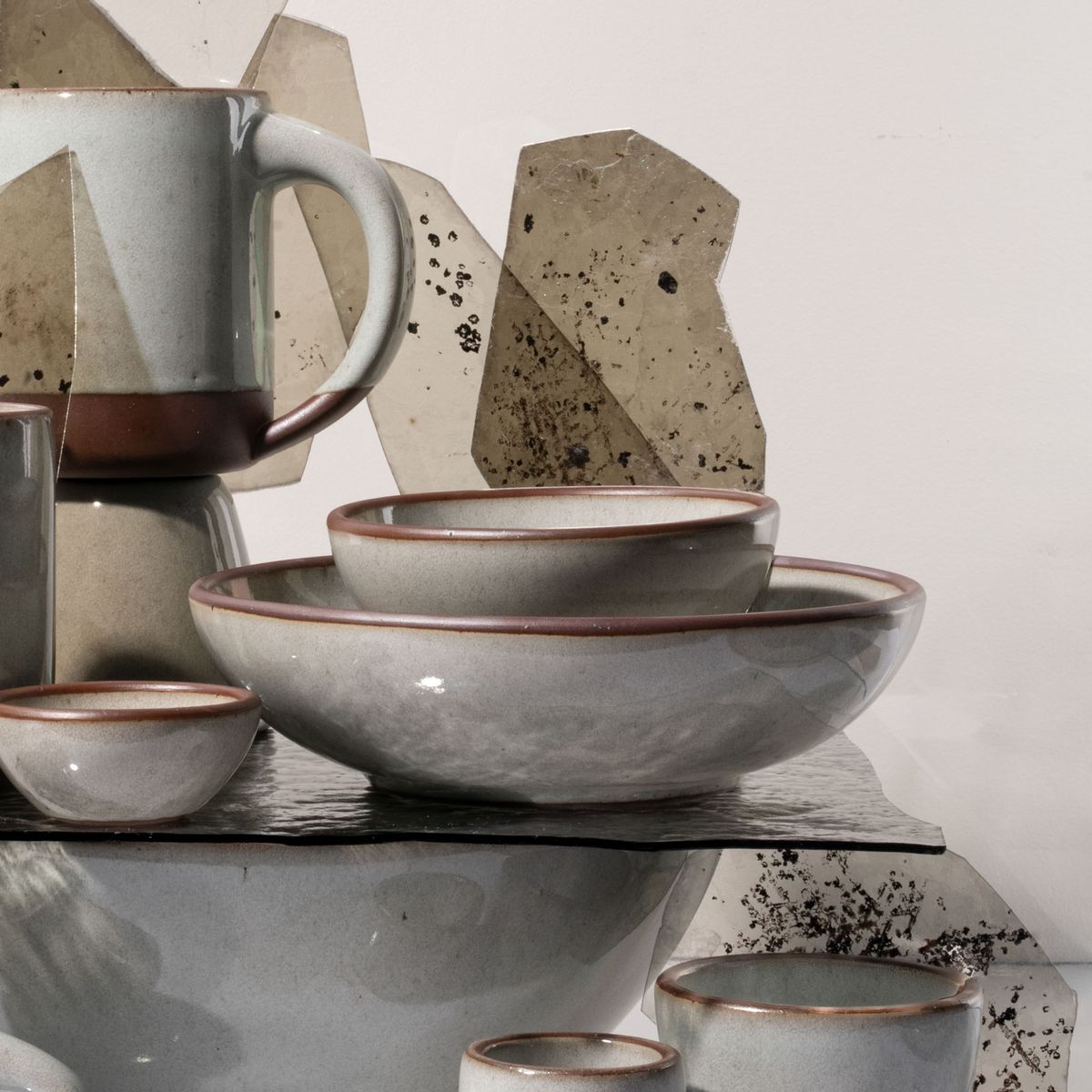 Stacked ceramic bowls and mugs in a reflective icy white glaze with brown rims, arranged with textured glass panels against a neutral background.