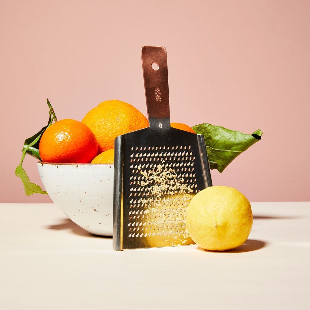 An East Fork bowl containing citrus fruit with green leaves and a lemon sit near a rectangular metal grater that has pieces of lemon peel on it with a copper-colored handle