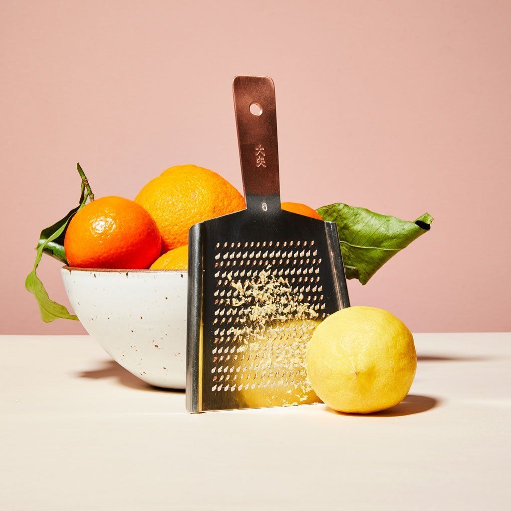 Citrus fruit with green leaves, an Eggshell East Fork bowl and a lemon sit near a rectangular metal grater that has pieces of lemon peel on it with a copper-colored handle