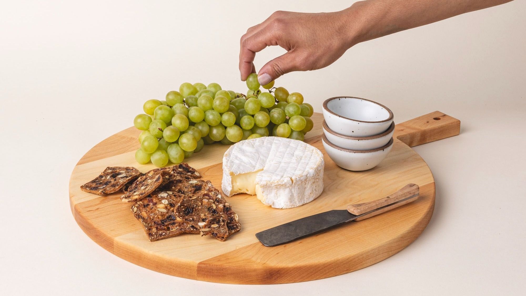A simple charcuterie board with cheese, crackers, and green grapes is on a round wooden serving board with a short rectangular handle with a hole for hanging