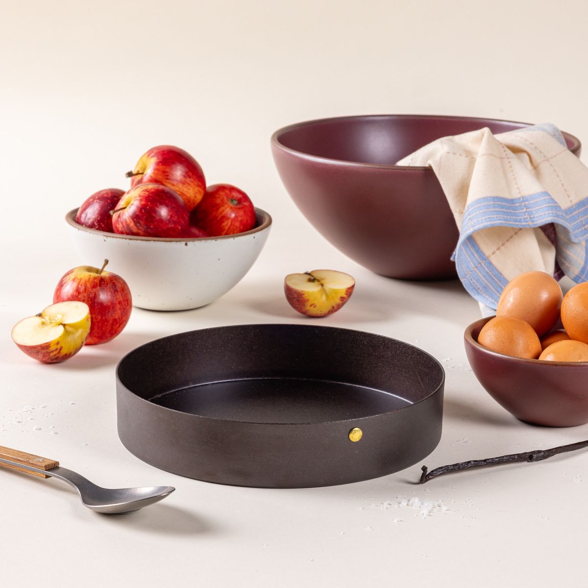 A round black metal cake pan, surrounded by large ceramic bowls, apples, and eggs.