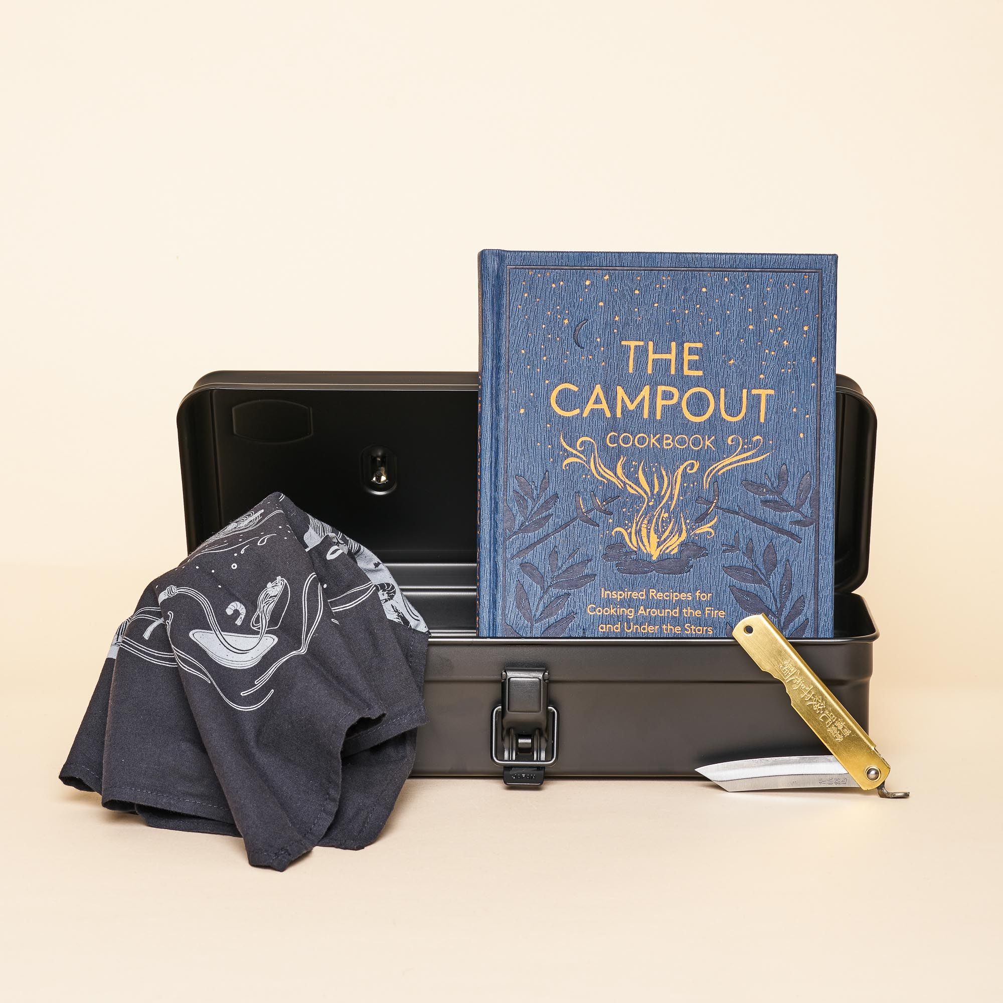 A steel box opens to display a navy blue cookbook ("The Campout Cookbook"). A hand holds a brass handled pocket knife with Japanese inscriptions on the handle. A black and white bandana sits in front of the box.