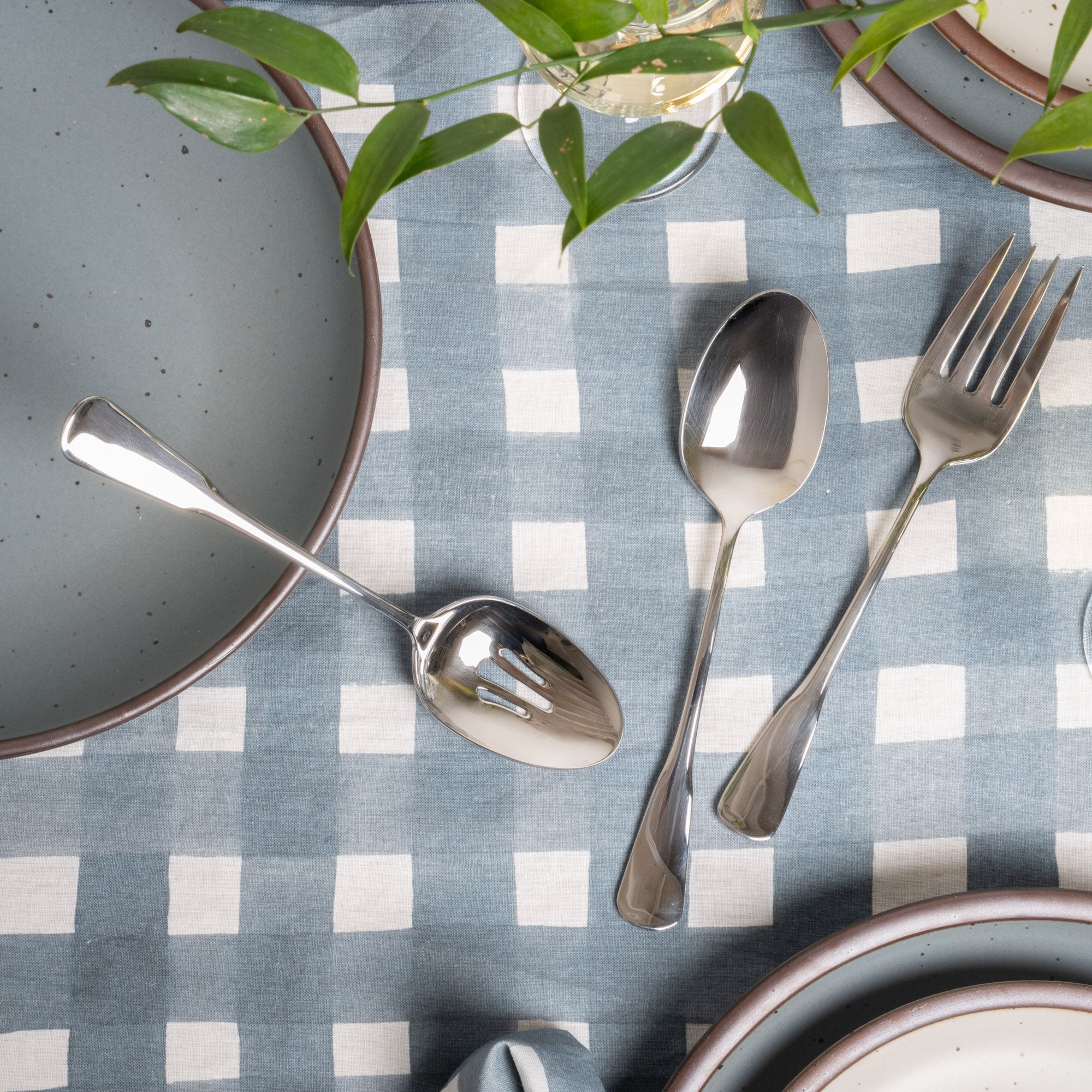 Three stainless steel serving utensils including a serving fork, serving spoon, and pierced spoon in a polished finish with a classic design, sitting on a blue-grey crosshatch tablecloth