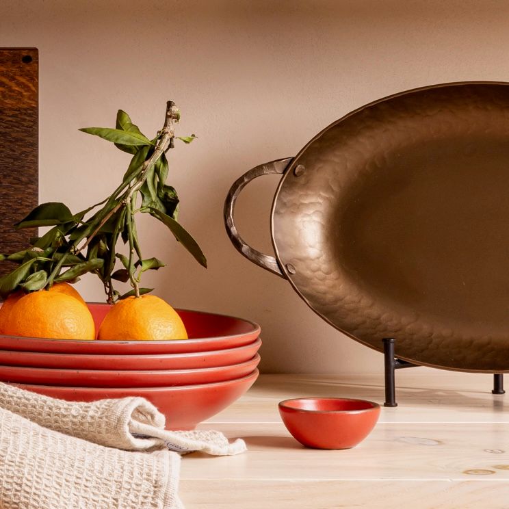 On a shelf sits a hammered oven roaster pan propped up, along with a stack of red ceramic coupe plates with oranges on top, a tiny red bowl, and a natural kitchen towel.