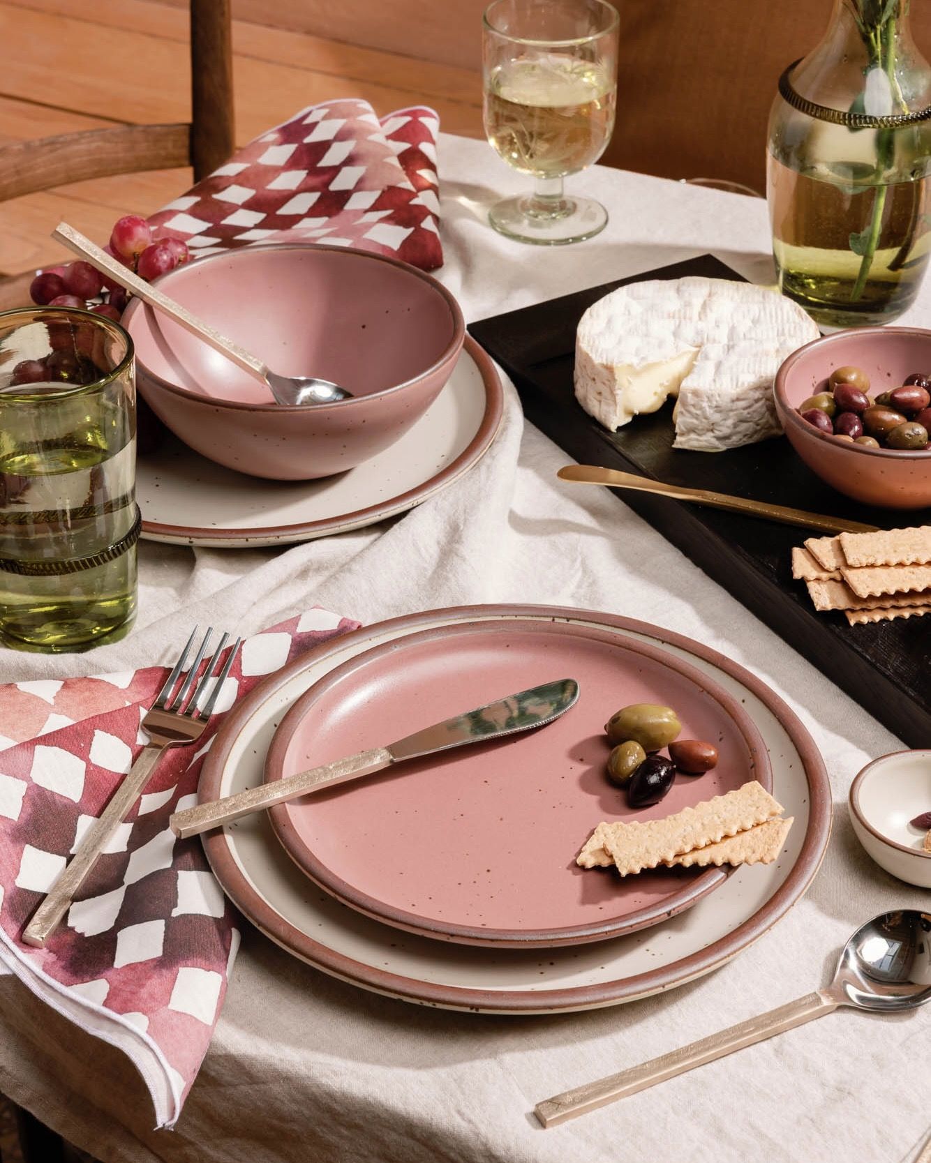 Table setting with ceramic plates in dusty mauve pink, brass flatware, and watercolor diamond napkins, and a charcuterie board.