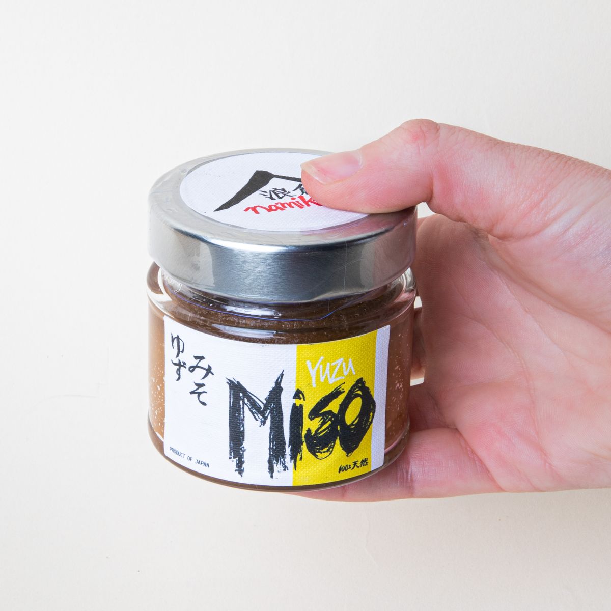 A hand holding a small clear jar with a metal screw cap with a white and yellow label that reads "Miso".