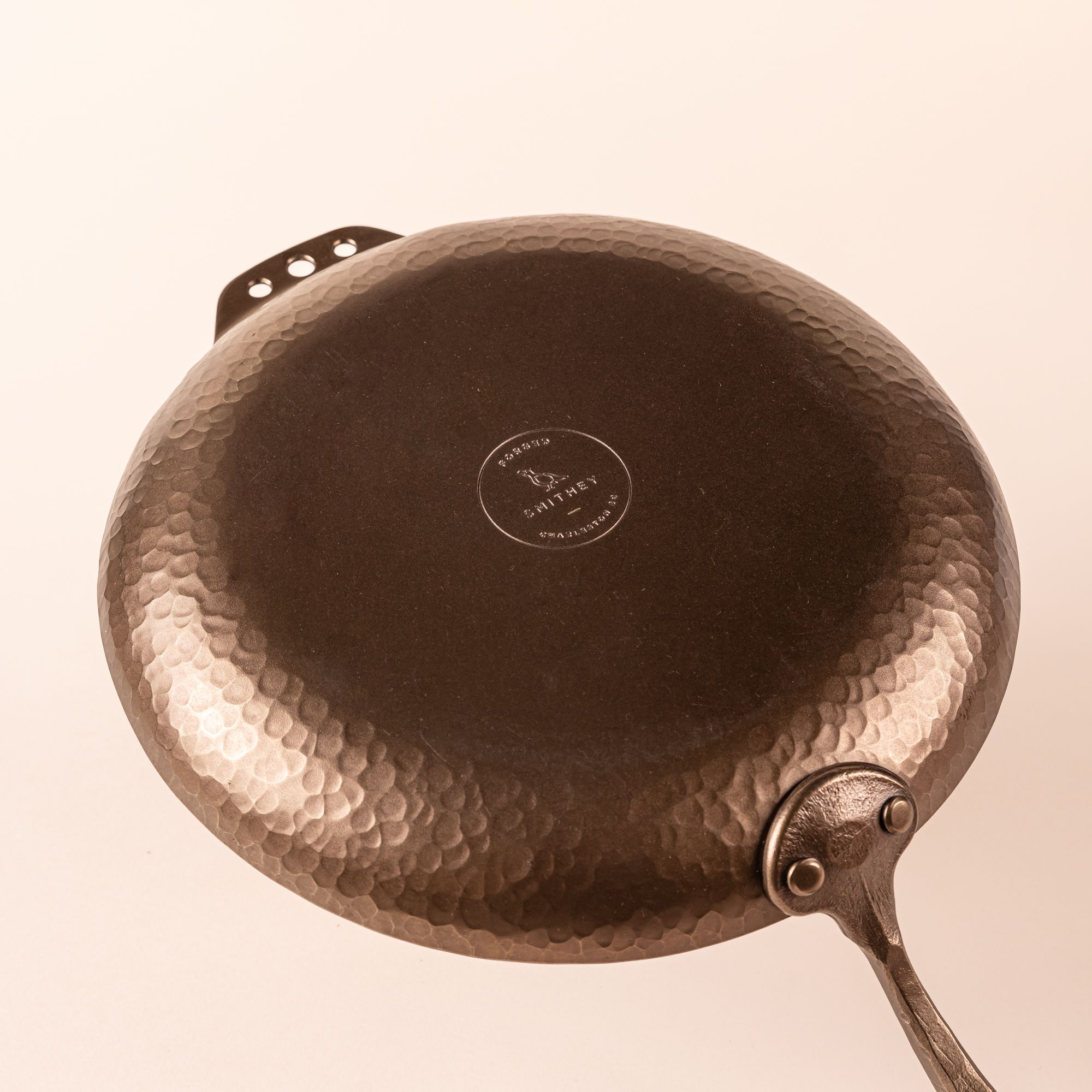 The bottom of a carbon steel farmhouse skillet with a hammered texture and hand forged design. The bottom features a stamp for the brand, Smithey.