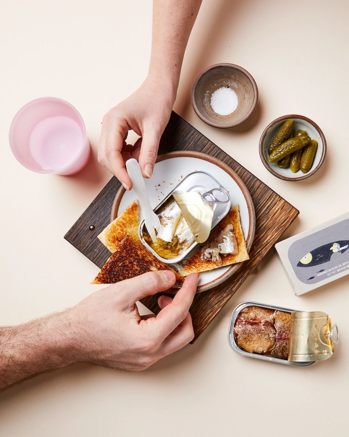 Two hands reaching into a wooden cutting board with assorted tins of fish