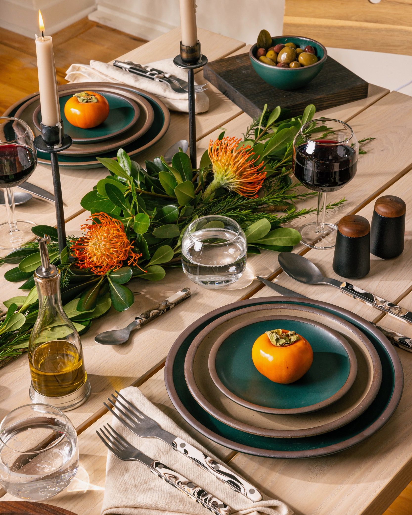 A tablescape featuring ceramic pottery in deep teal and neutral colors, along with a greenery centerpiece, wine glasses, sophisticated flatware, and lit taper candles.