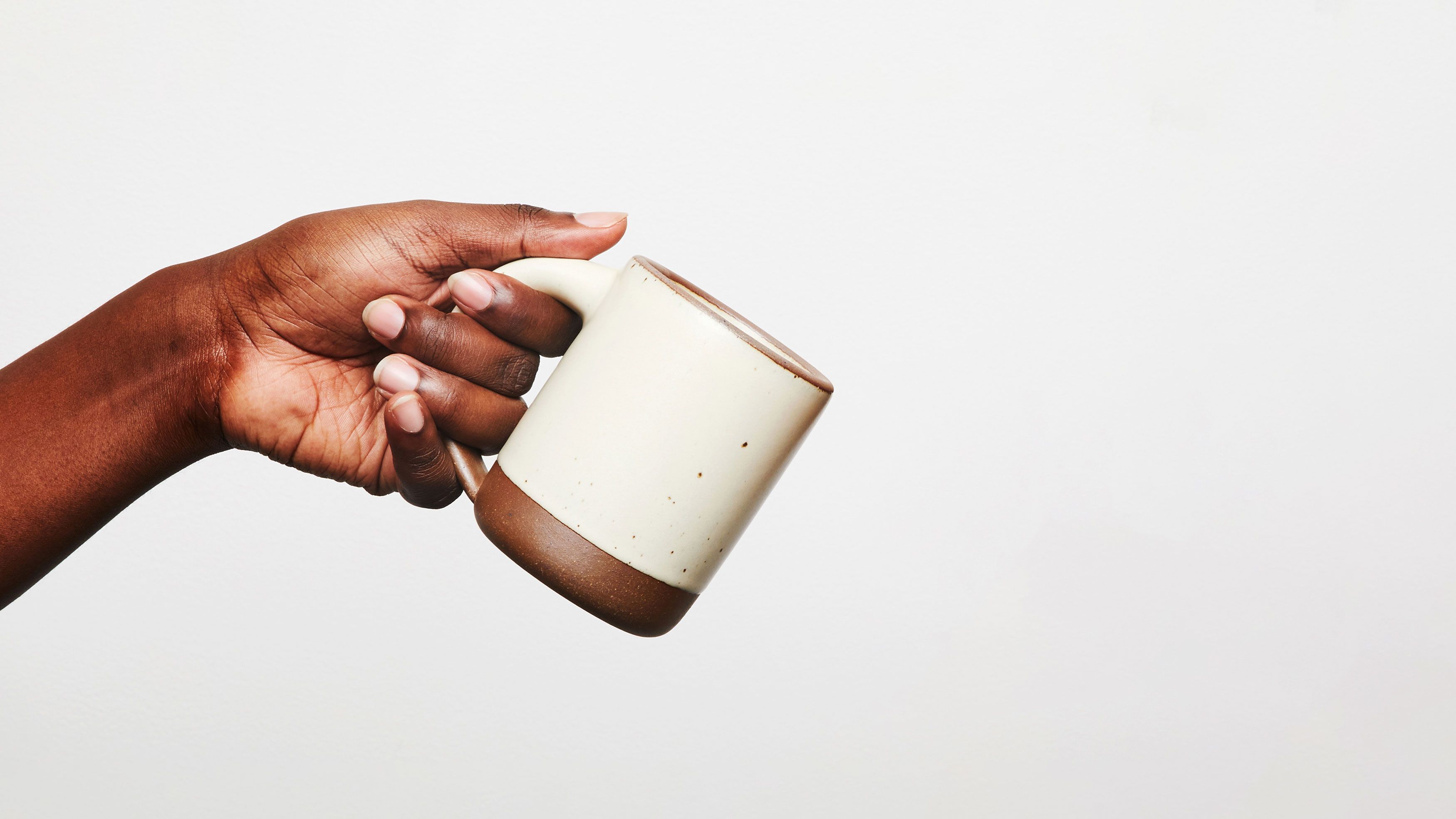 A hand holds an cool, white ceramic mug with a raw clay rim, with three fingers fitting effortlessly through the mugs handle