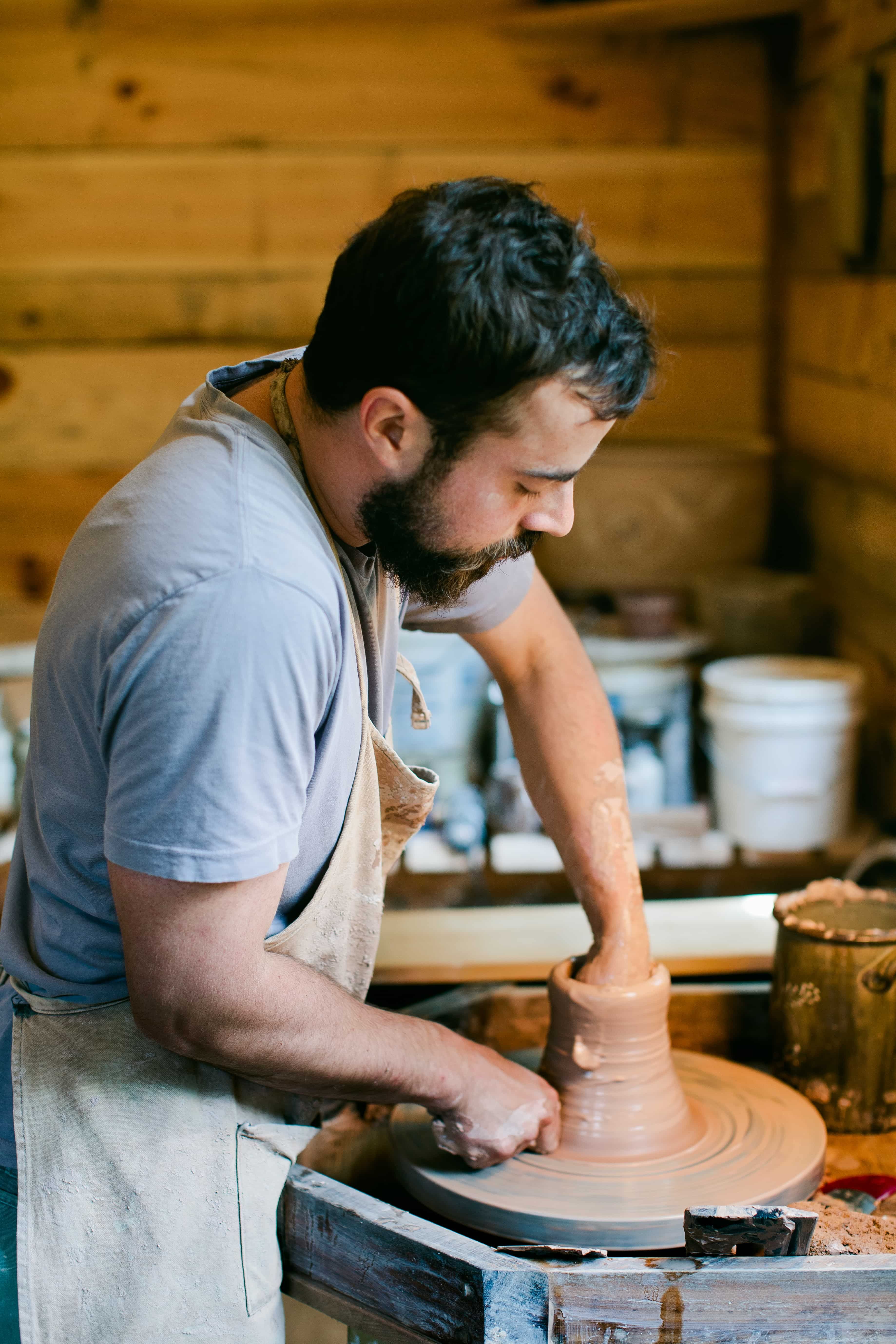 Alex Matisse is in a potter's workshop in front of a potter's wheel and forming a piece of pottery