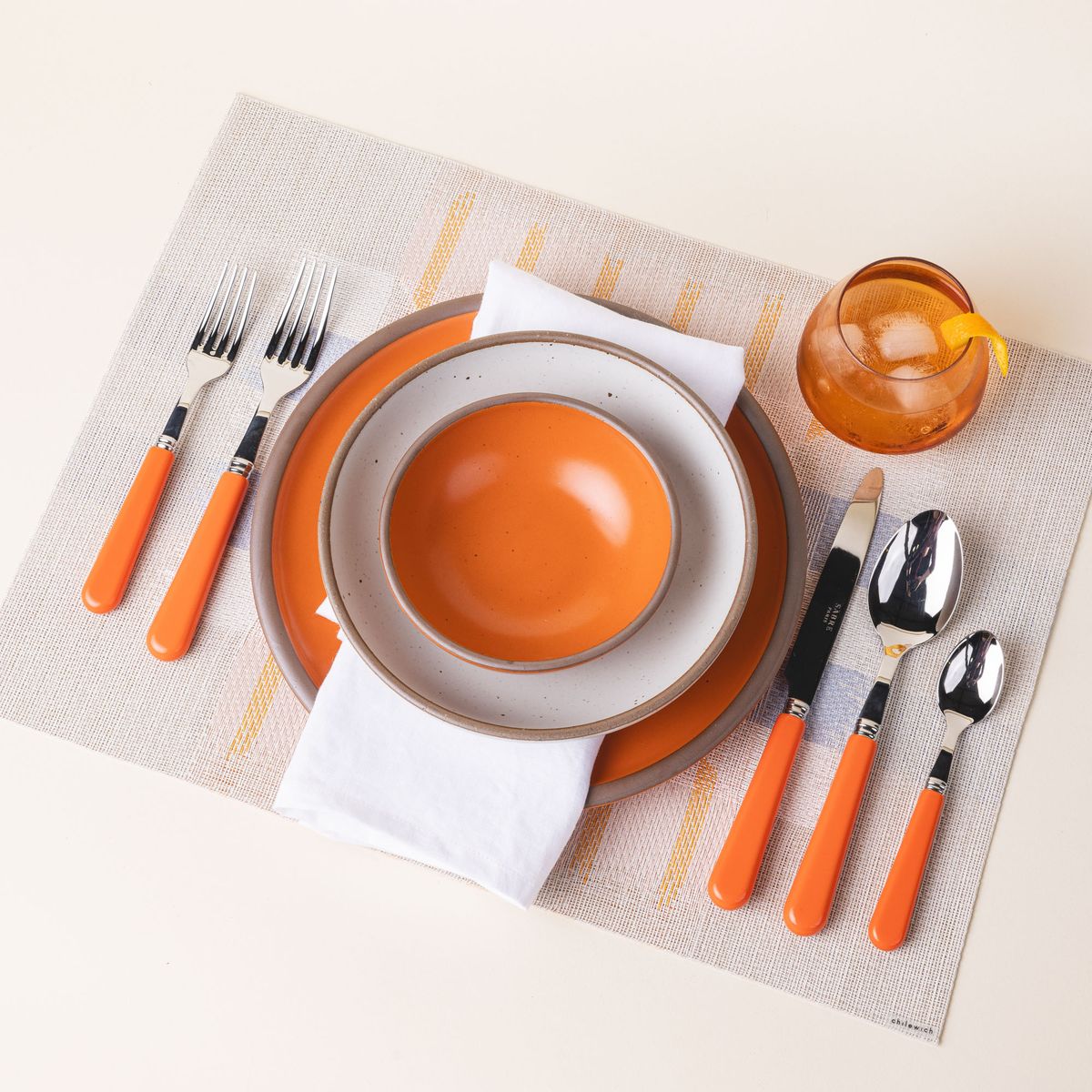 A place setting with a placemat with a soft artful geometric design featuring neutral, orange, white, and blue colors, ceramic bold orange bowls and plates, orange flatware, and an orange whiskey snifter.
