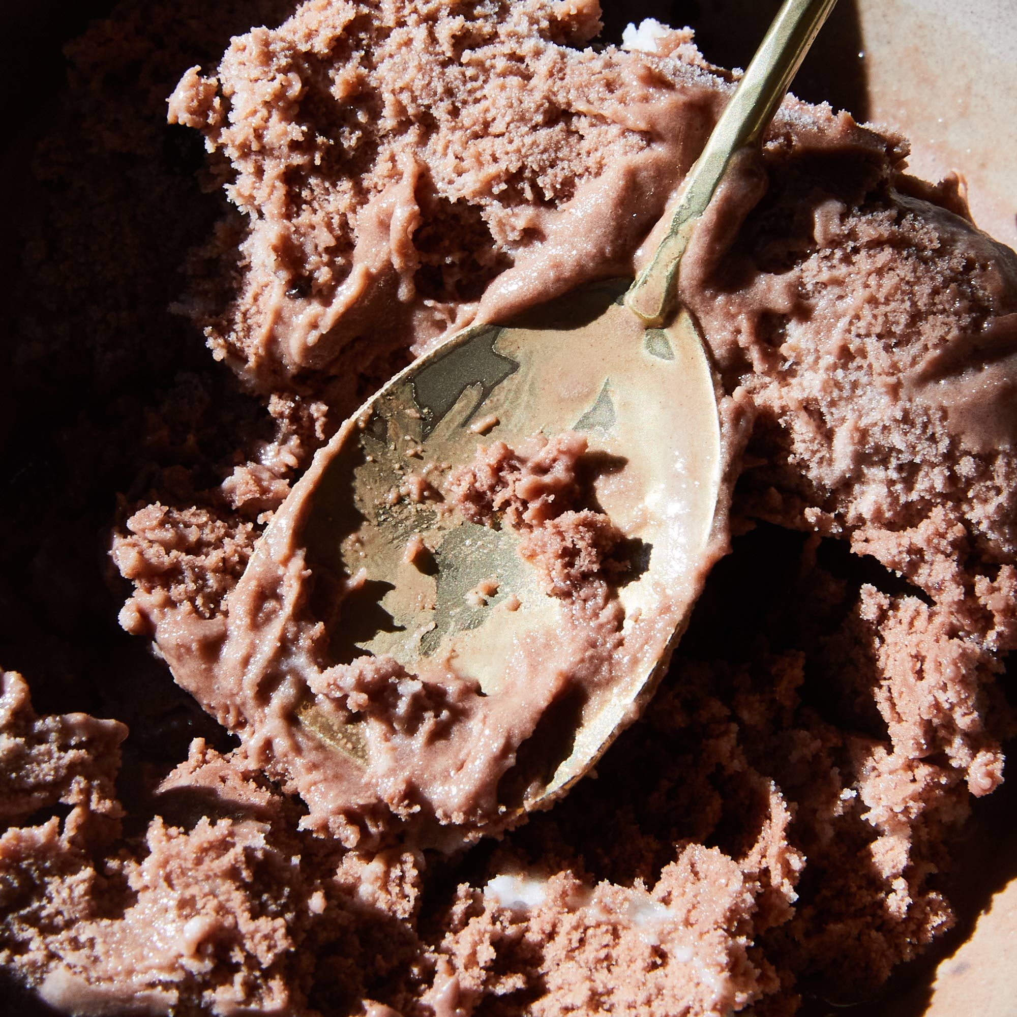 A scoop of chocolate ice cream with a brass spoon digging into it, with some ice cream on the bowl