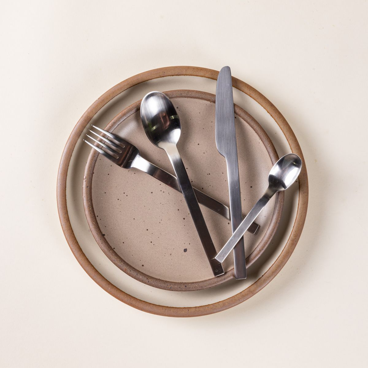 A simple steel fork, knife, spoon, and teaspoon lay on 2 ceramic neutral plates.